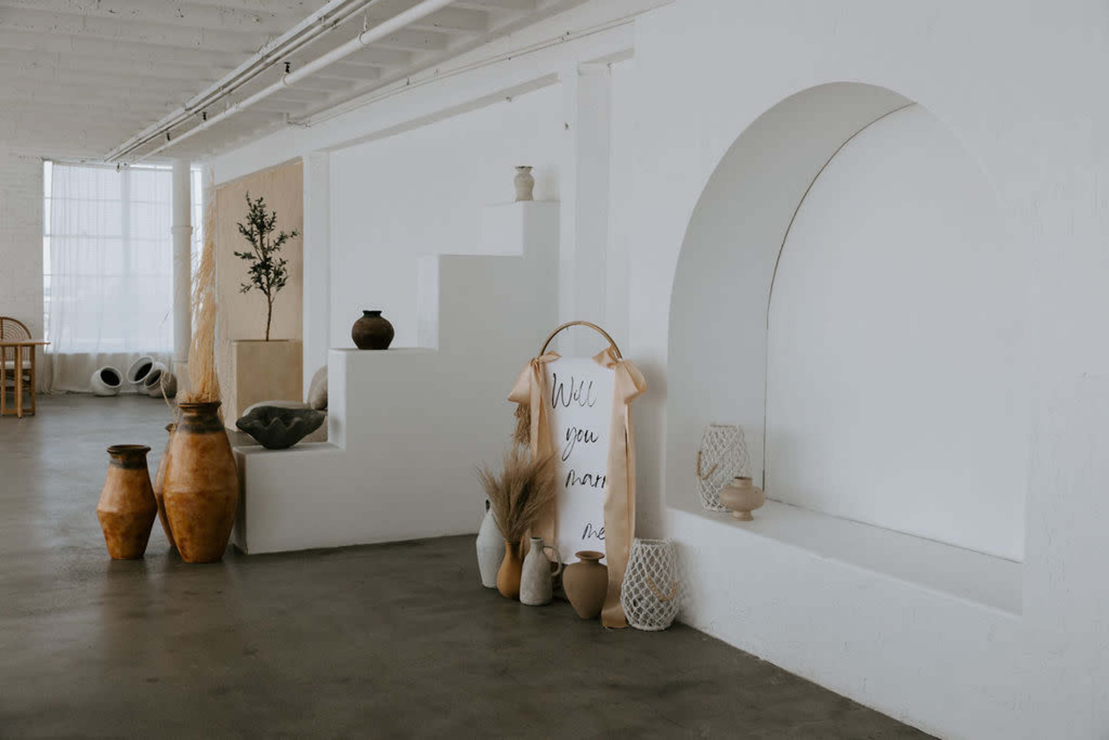 A minimalist interior space featuring white walls, a series of steps, decorative pottery, and a wall hanging with text.
