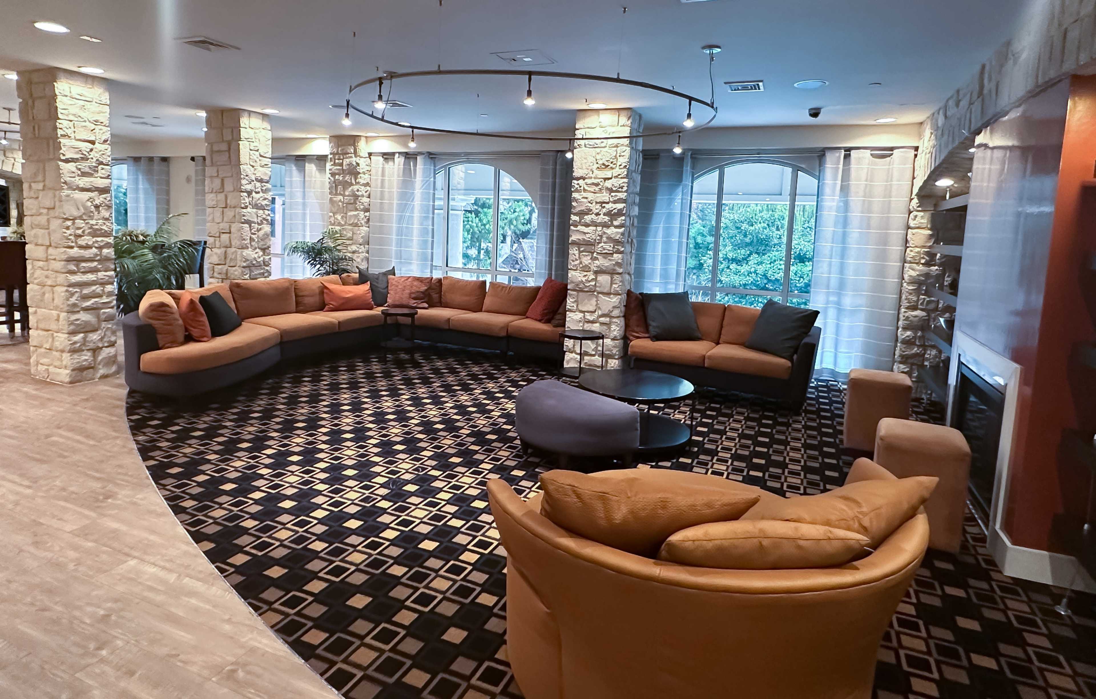 A spacious lounge area with a circular seating arrangement featuring various couches, a coffee table, and large windows overlooking greenery.