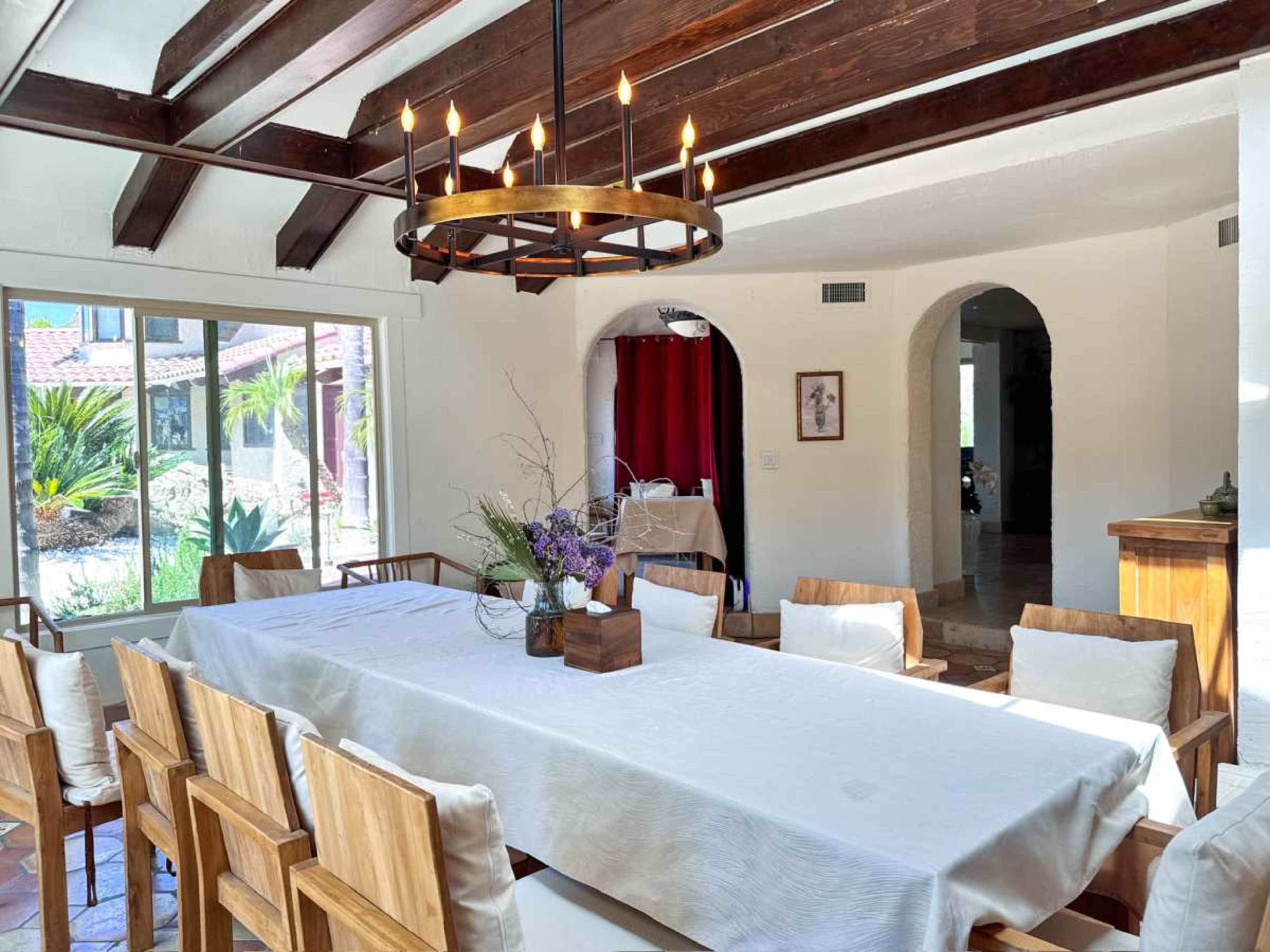 A long wooden dining table is set in a sunlit room with exposed beams, surrounded by chairs and adorned with a centerpiece of flowers.