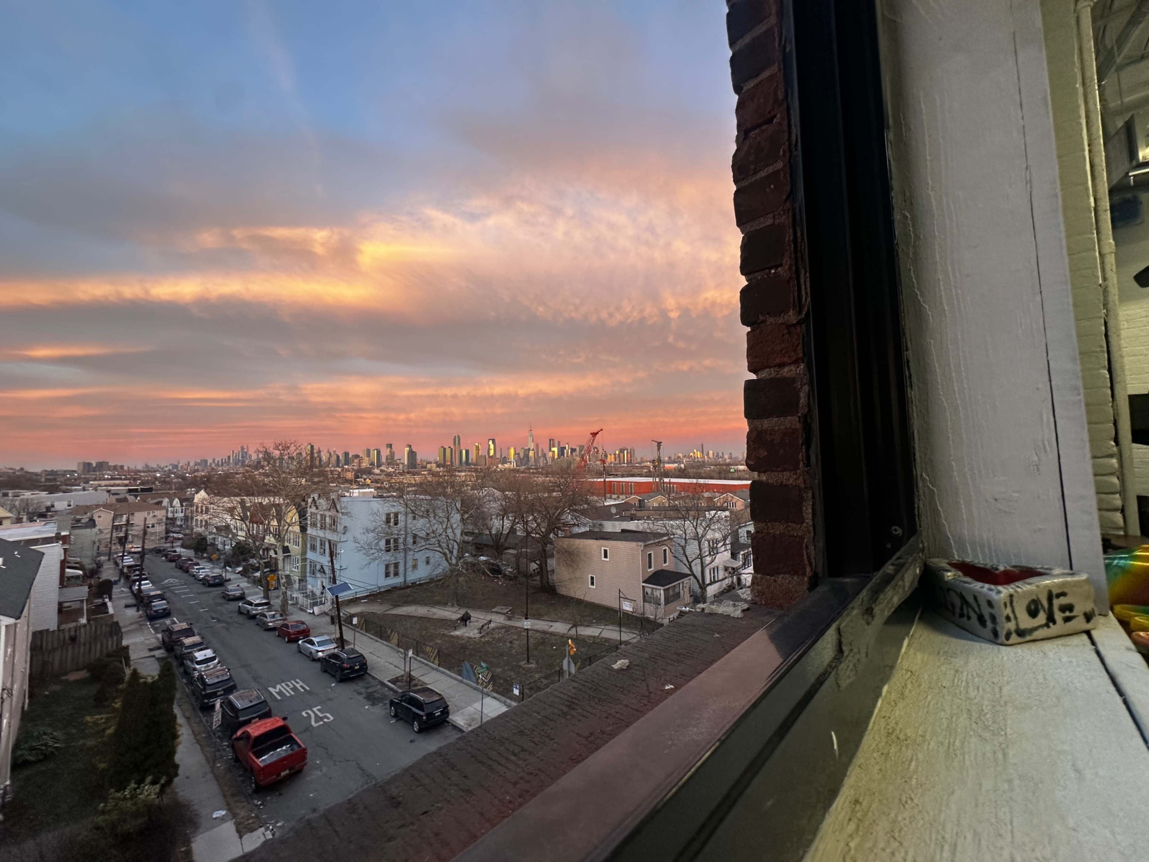 A view of a city skyline at sunset is framed by a window, with cars parked on a residential street below.