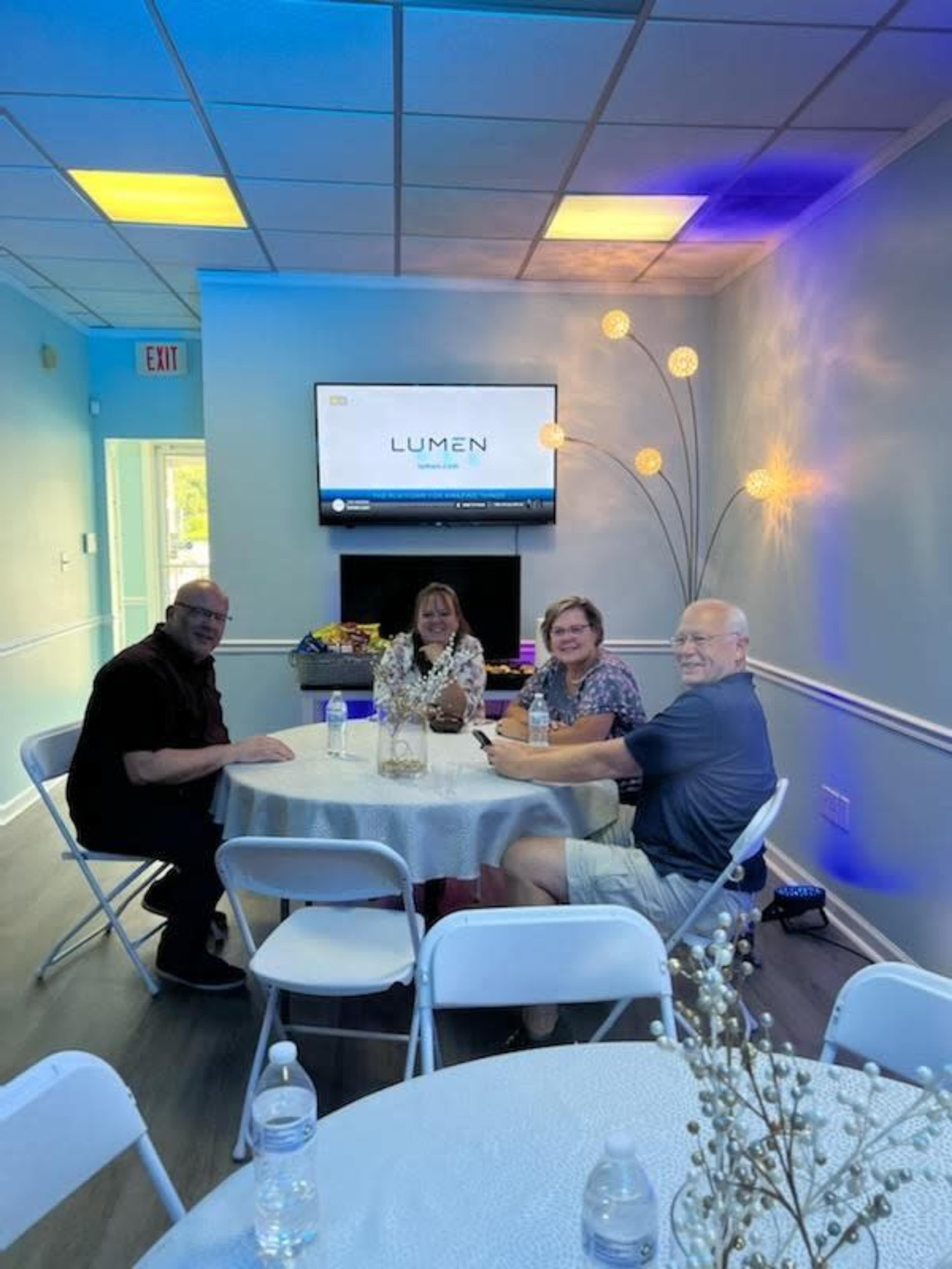 Four people sit at a table with water bottles in a brightly lit room featuring a wall-mounted television and decorative lighting.