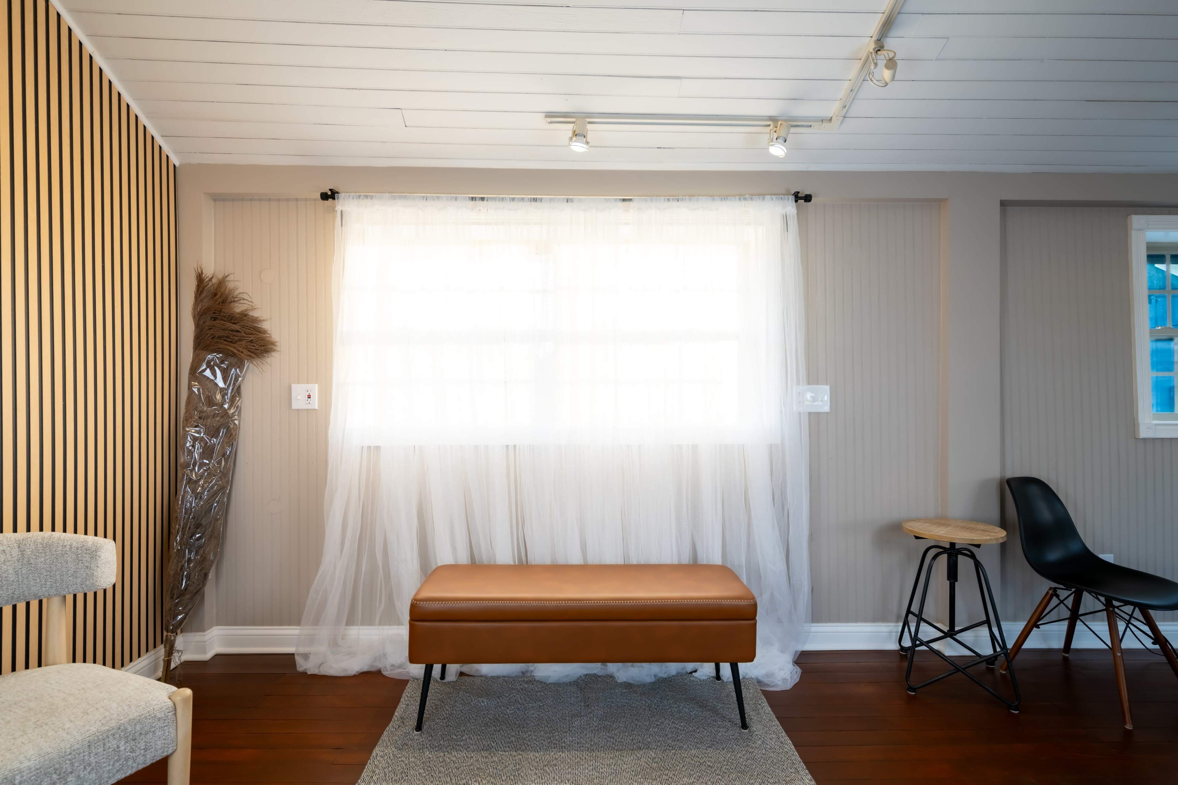 The image shows a minimalist interior space with a brown leather bench, a textured rug, and a sheer curtain covering a window.