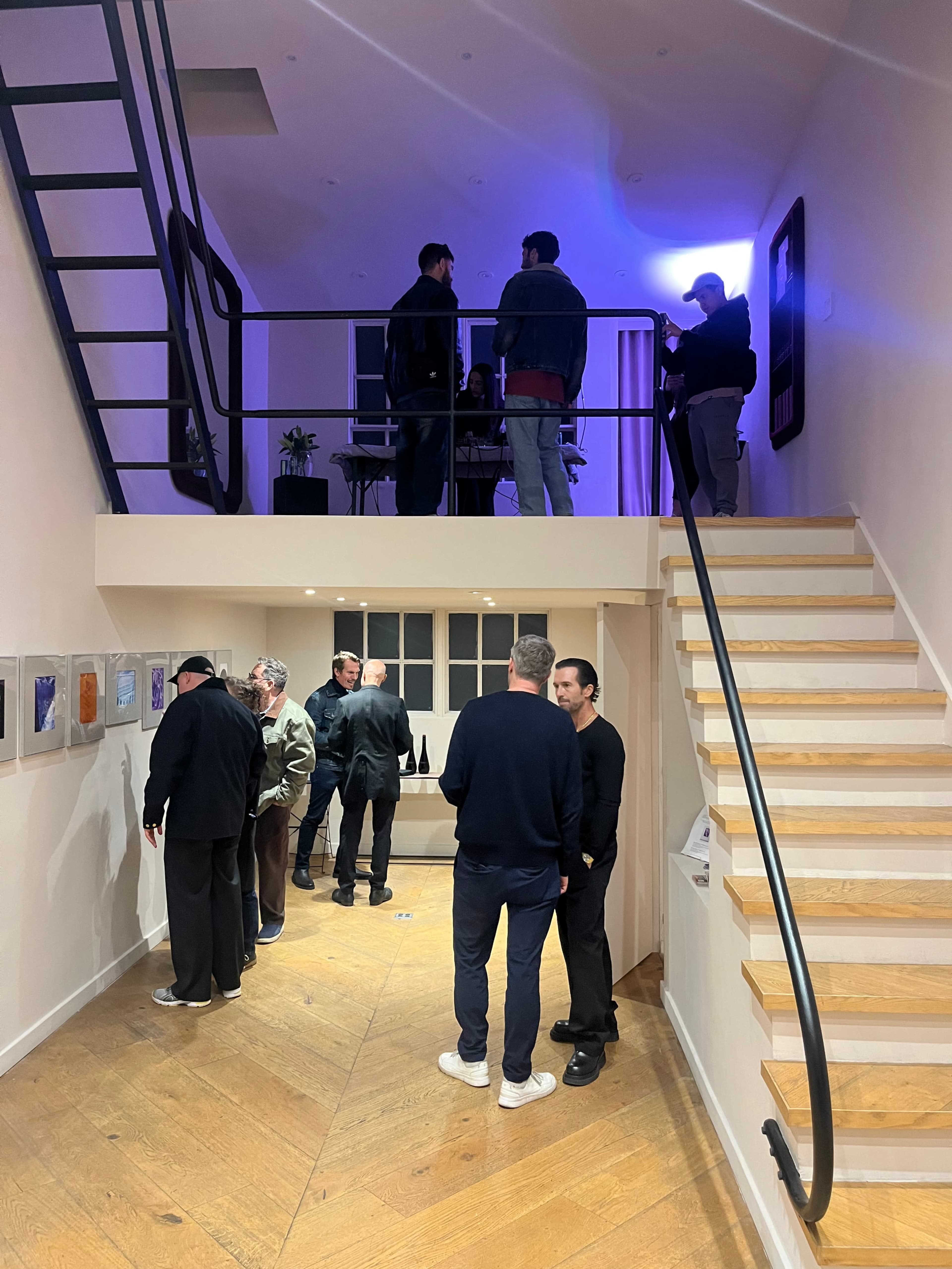 The image shows a contemporary gallery space with two levels, where several people are engaged in conversation and viewing art exhibits on the walls.