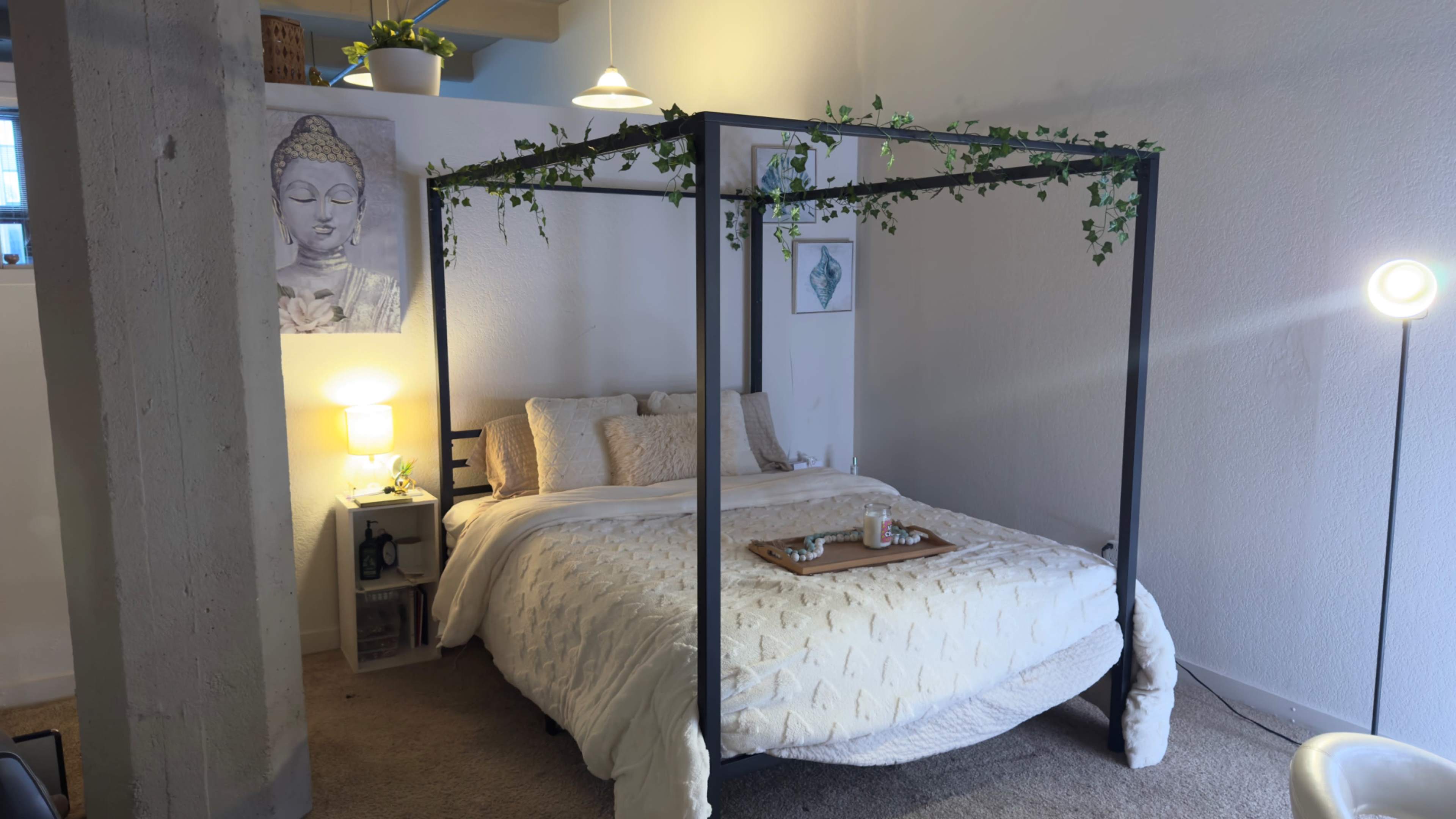 A bedroom featuring a four-poster bed with a white quilt, a bedside table with a lamp, and decor including a wall art piece of a Buddha and some greenery.