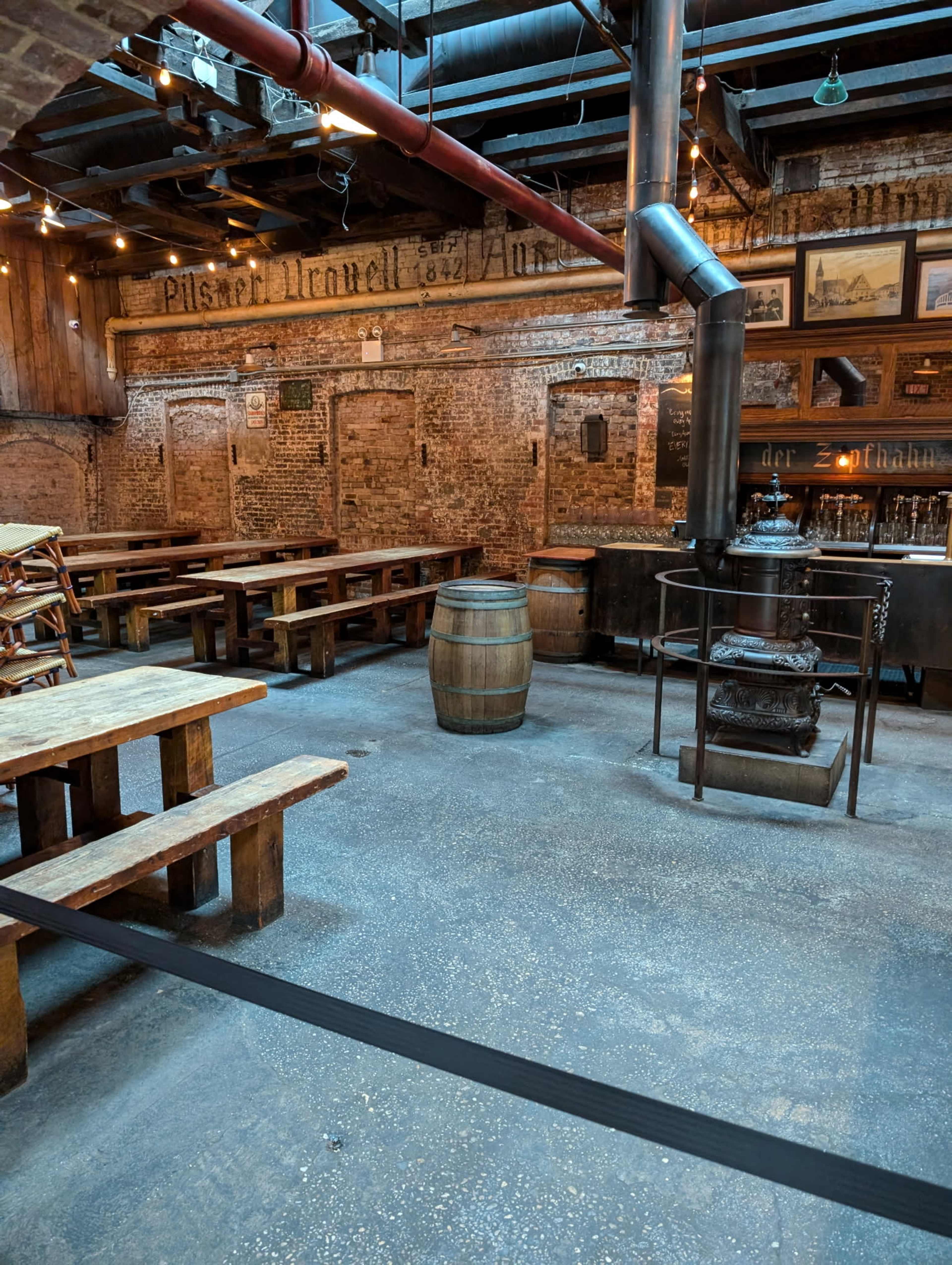 The image shows an industrial-style interior of a bar or restaurant featuring wooden benches, a barrel, and a vintage stove.