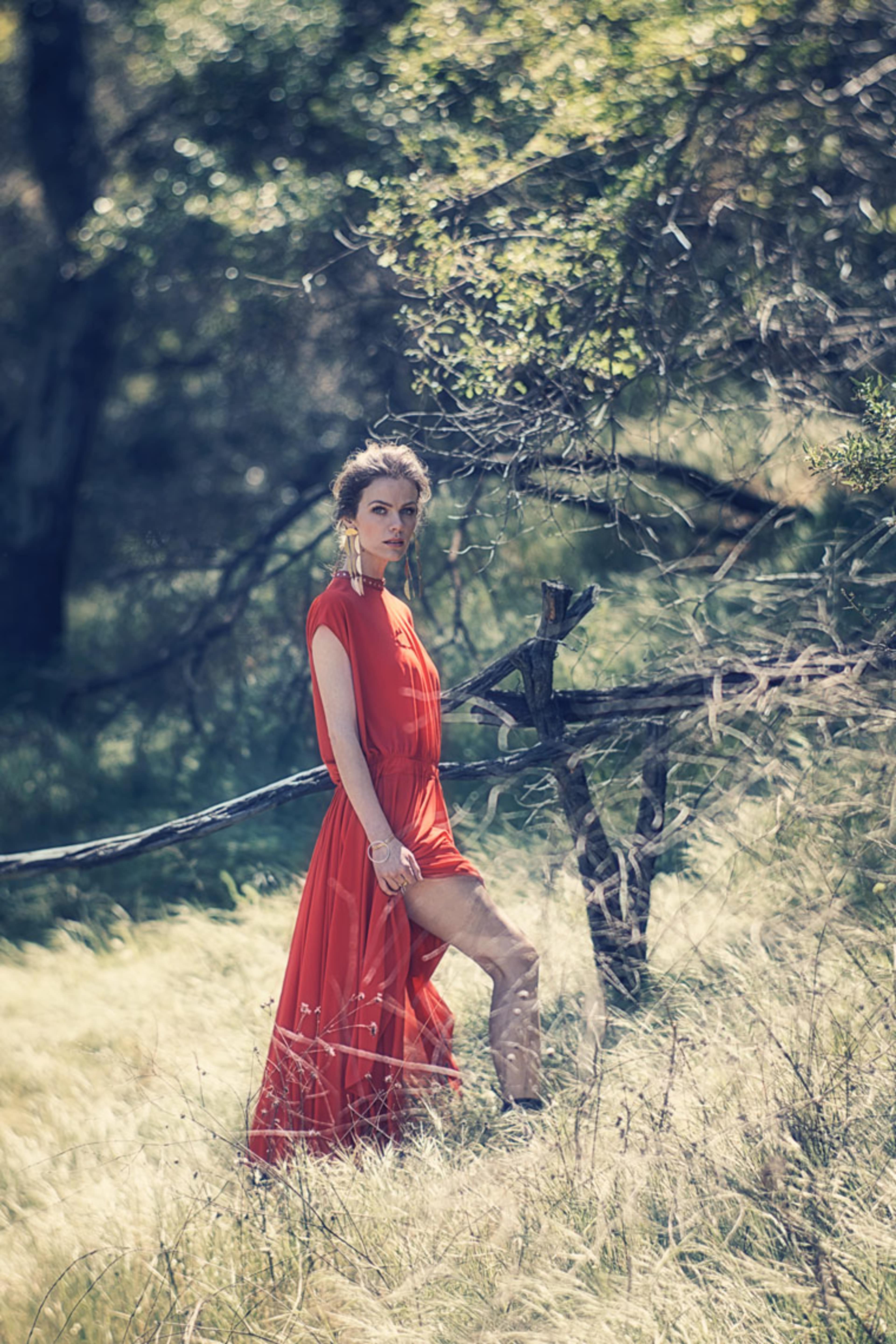A woman in a red dress stands among tall grass and trees in a natural setting.
