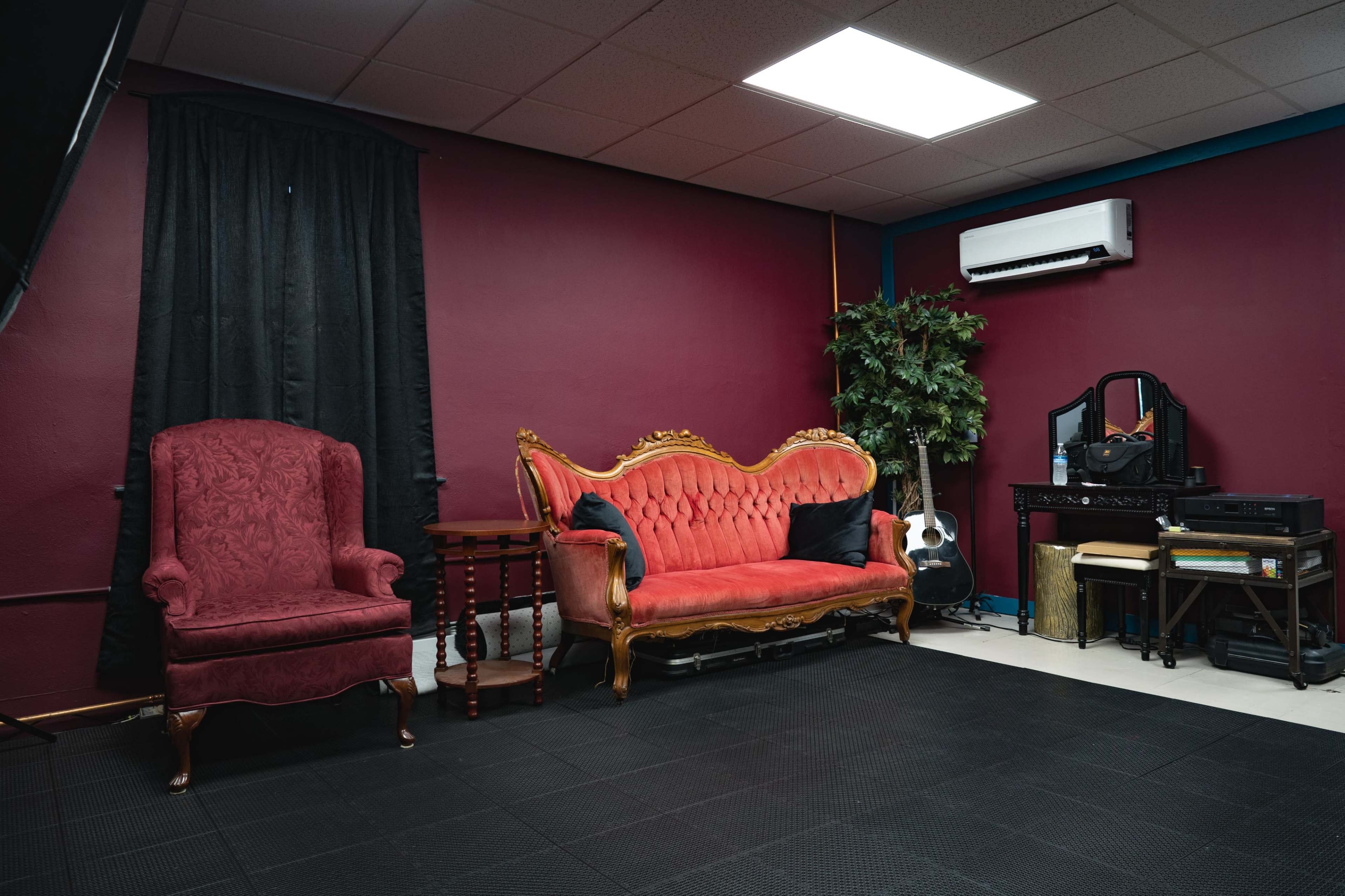 The image shows a room with a maroon wall featuring a red vintage couch, an armchair, a small wooden table, and a plant, along with various musical instruments and a mirror.