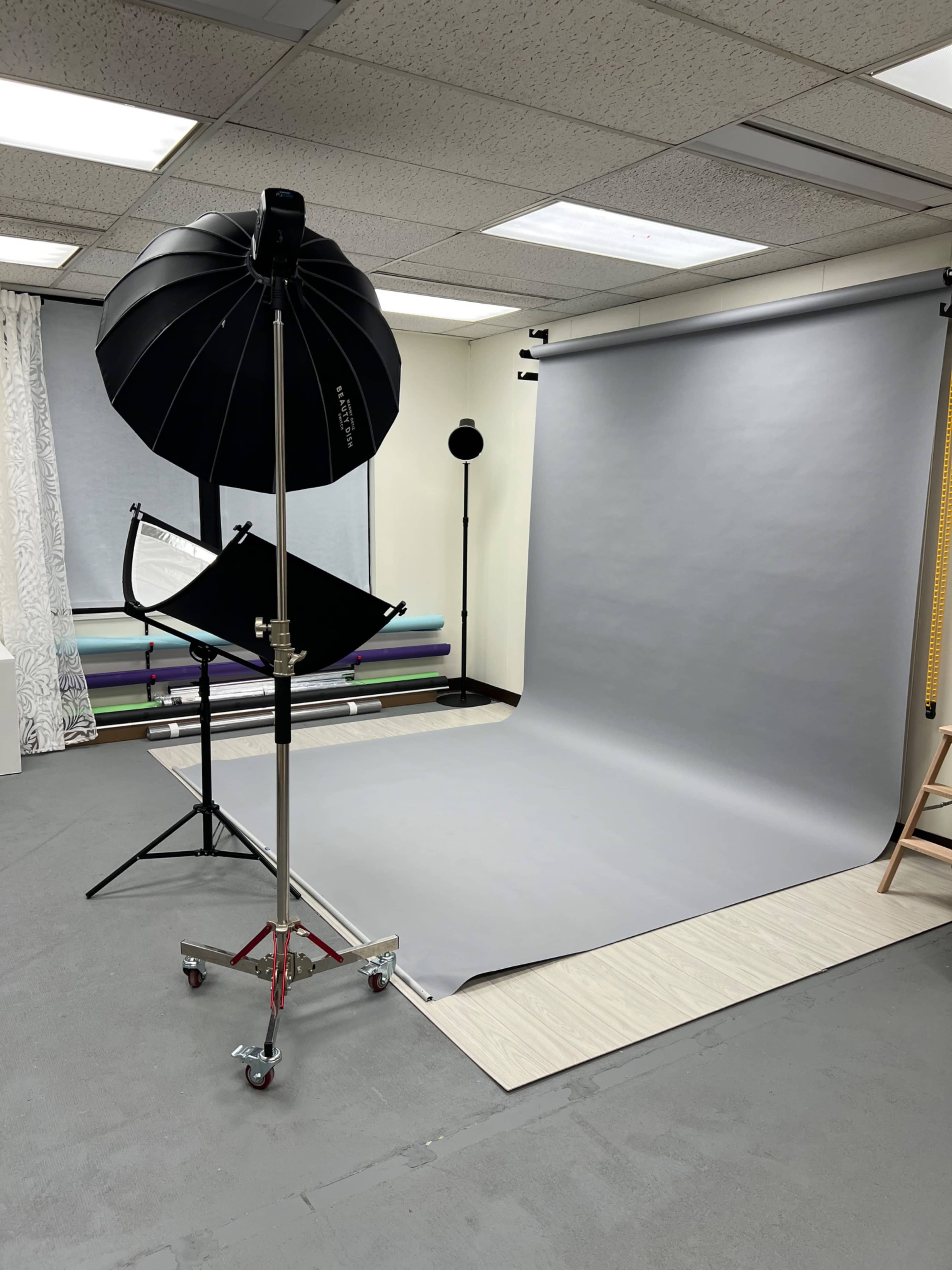 The image shows a photography setup featuring a gray backdrop, lighting equipment, and various colored backdrops in a studio space.