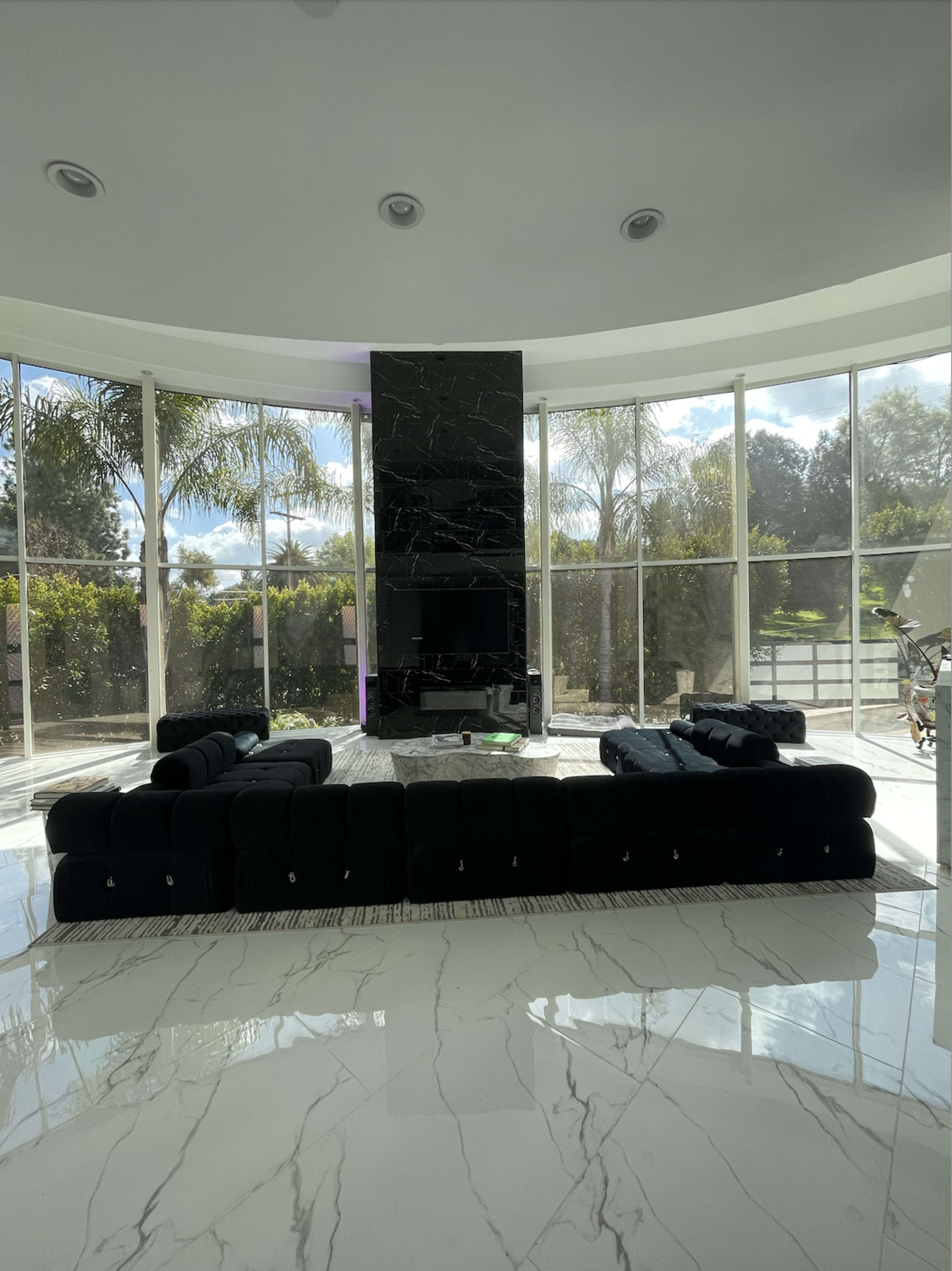 The image shows a modern living room featuring a black sectional sofa, a marble floor, large windows with views of greenery, and a black stone accent wall.