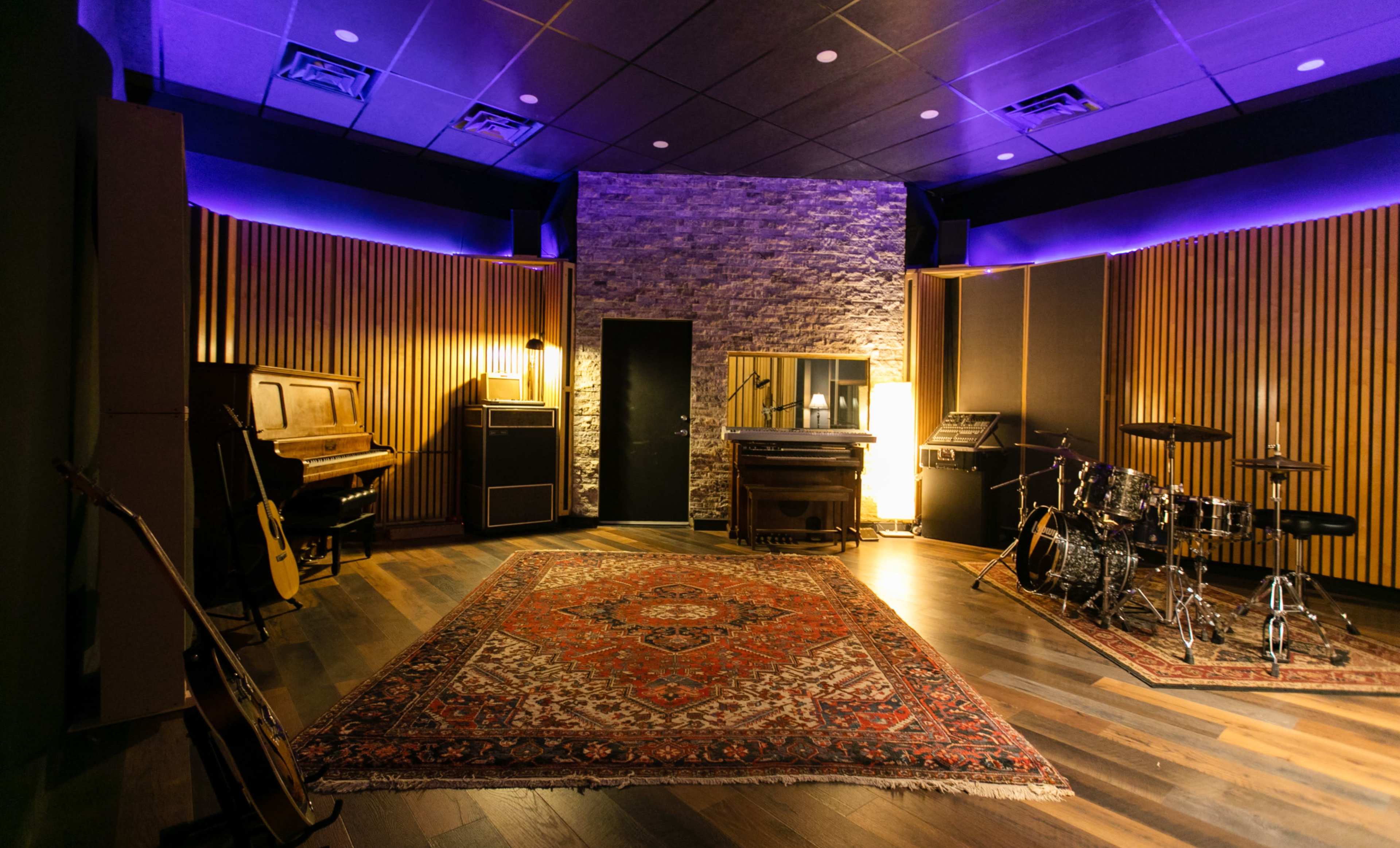 A music recording studio with wooden panels, a grand piano, a drum set, and various musical instruments arranged around a patterned rug.