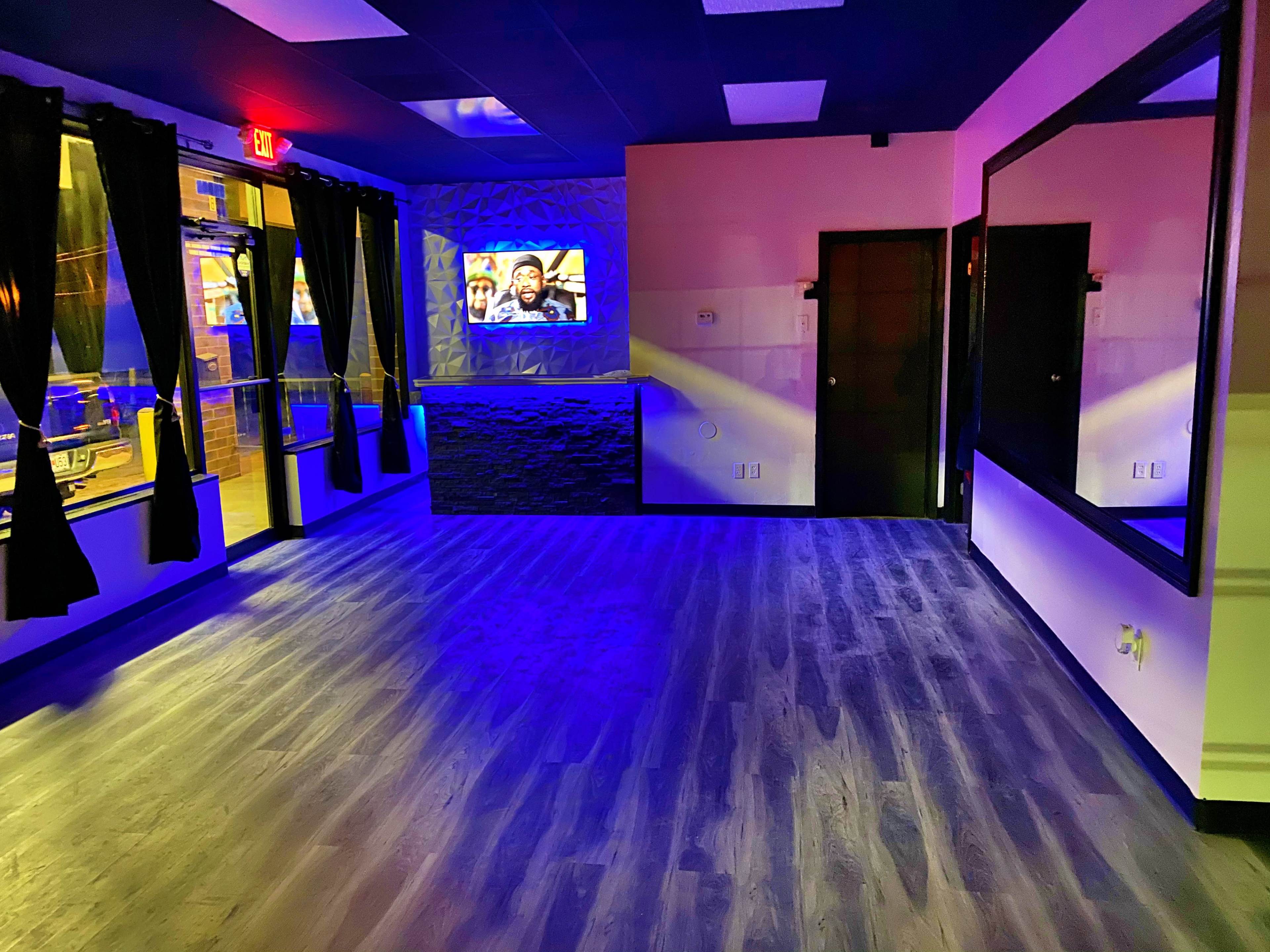 An empty room with blue lighting, featuring a textured wall, a television, large windows with dark curtains, and a mirror.