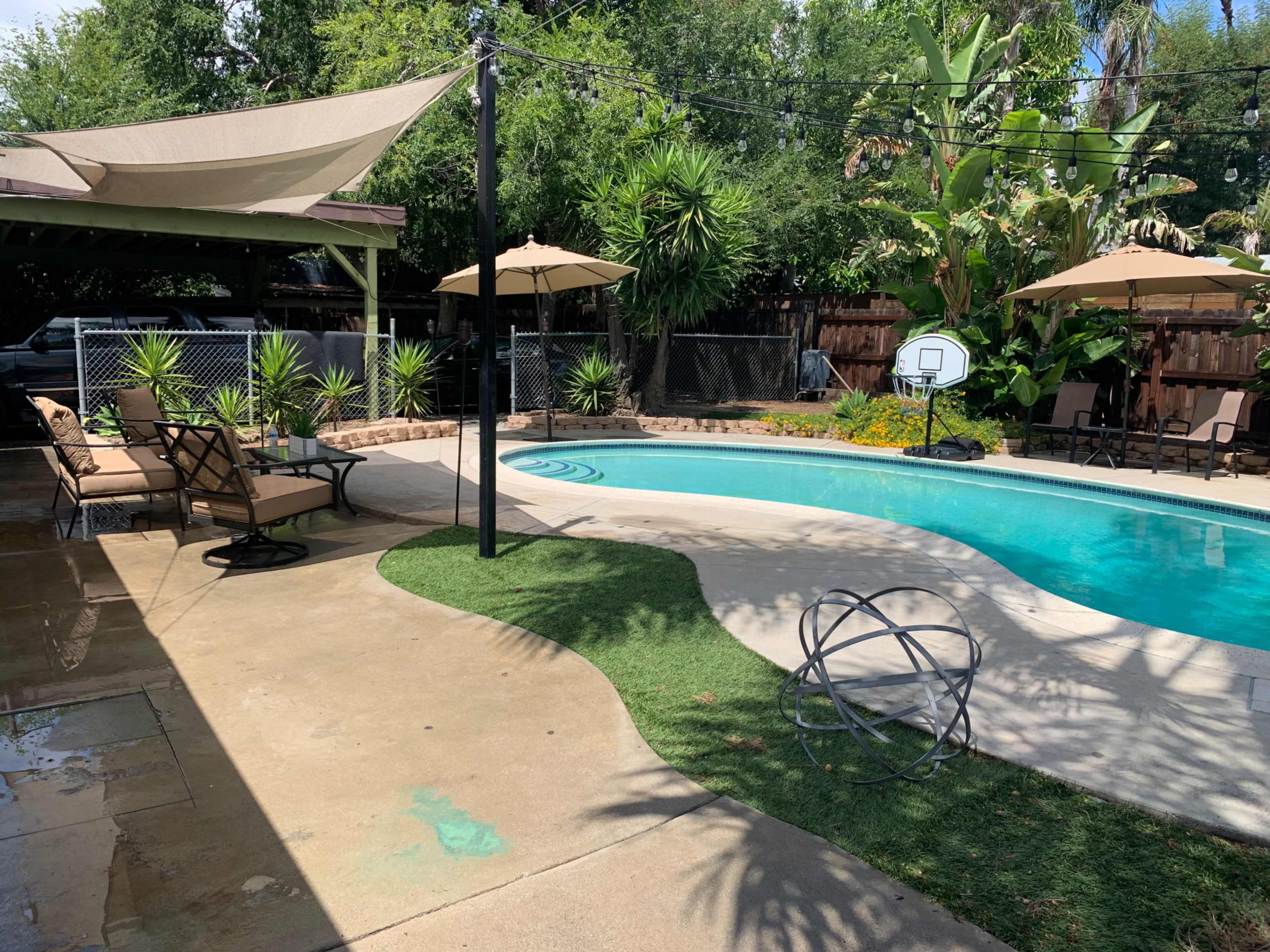 A backyard pool area with a swimming pool, lounge chairs, umbrellas, and landscaped greenery.