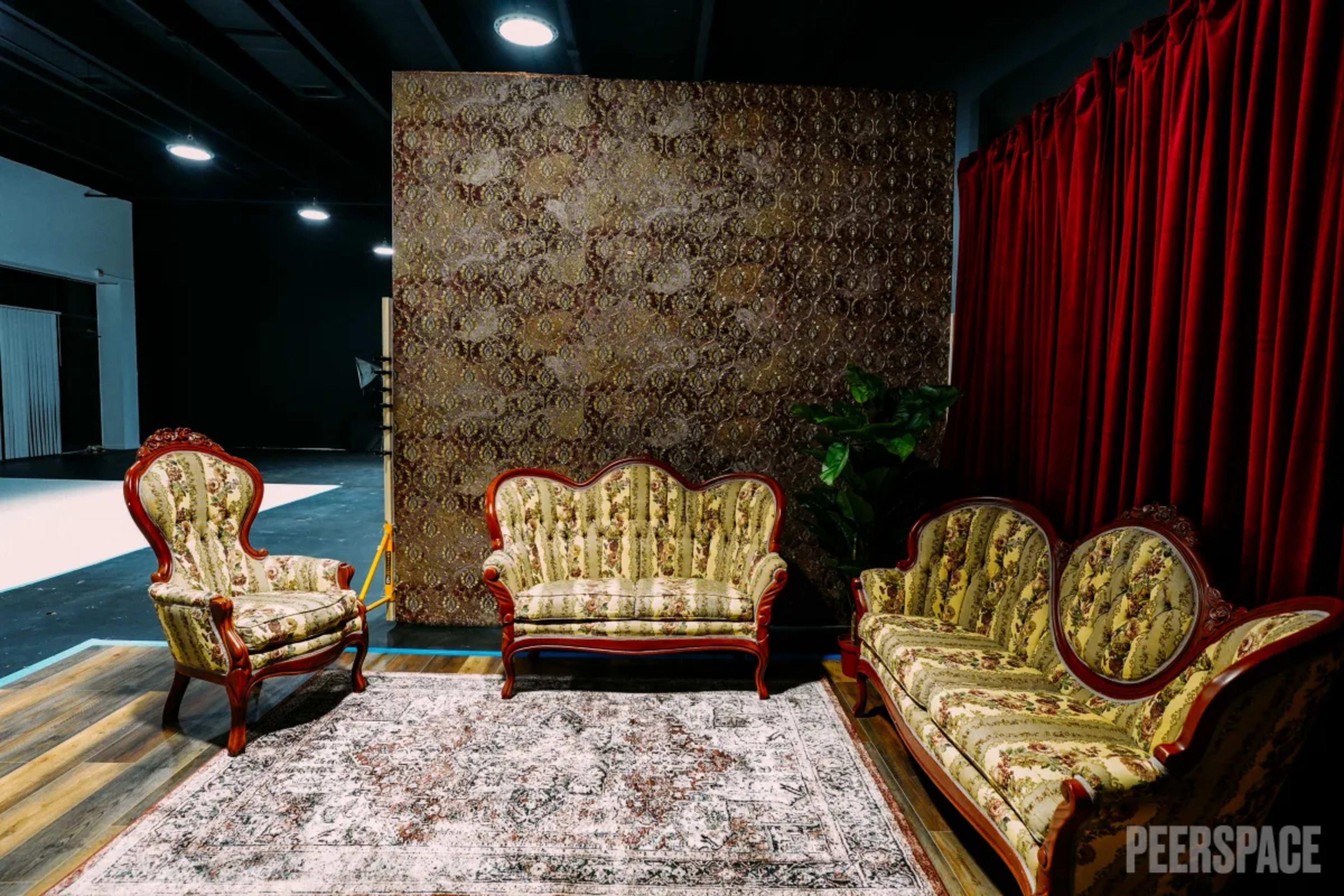The image shows a seating arrangement with ornate, patterned couches arranged in a well-lit room featuring a decorative wall and a red curtain.