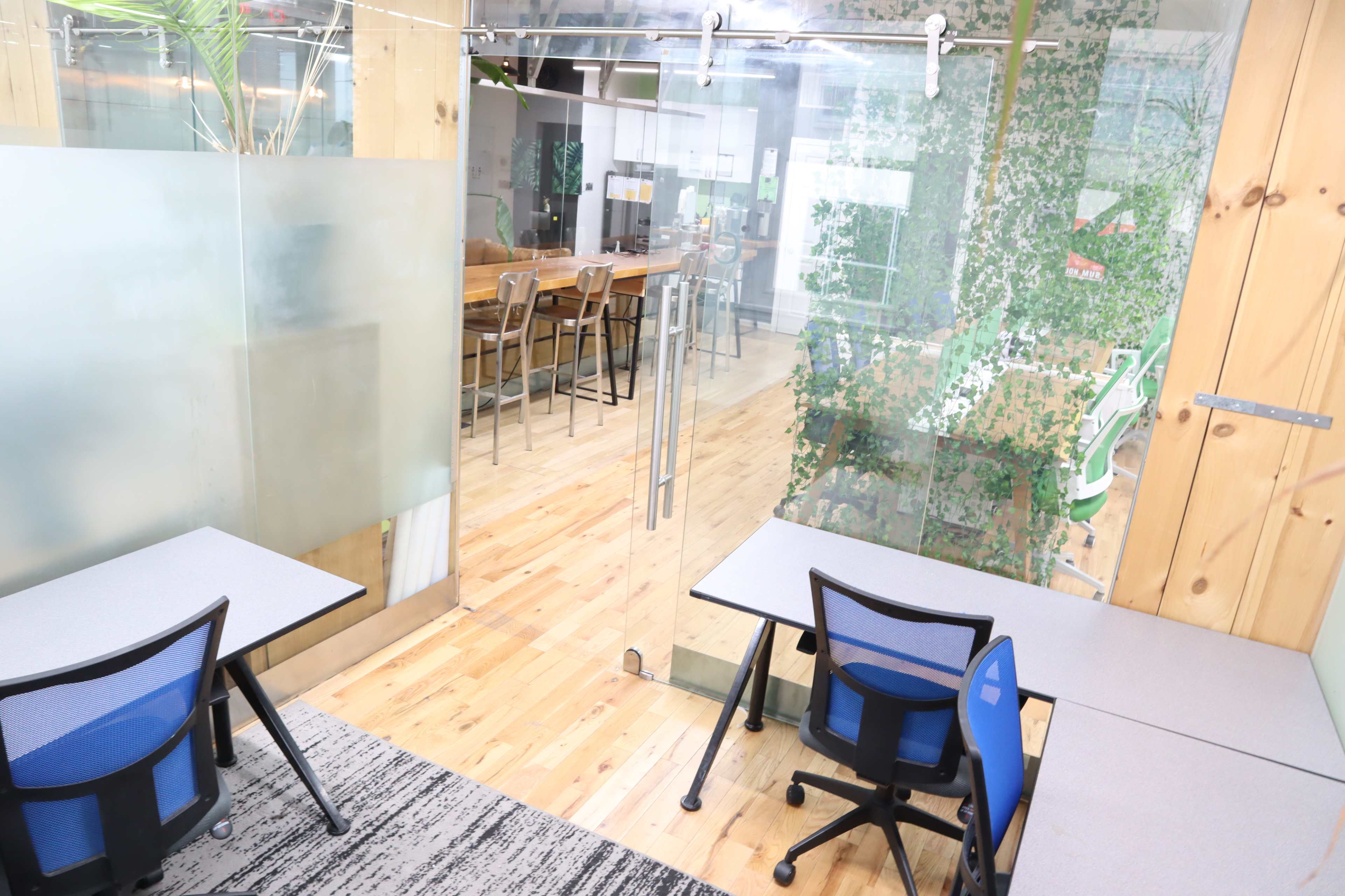 The image shows a modern office space with glass walls, featuring two small desks with chairs and a view of a wooden dining table and greenery beyond.