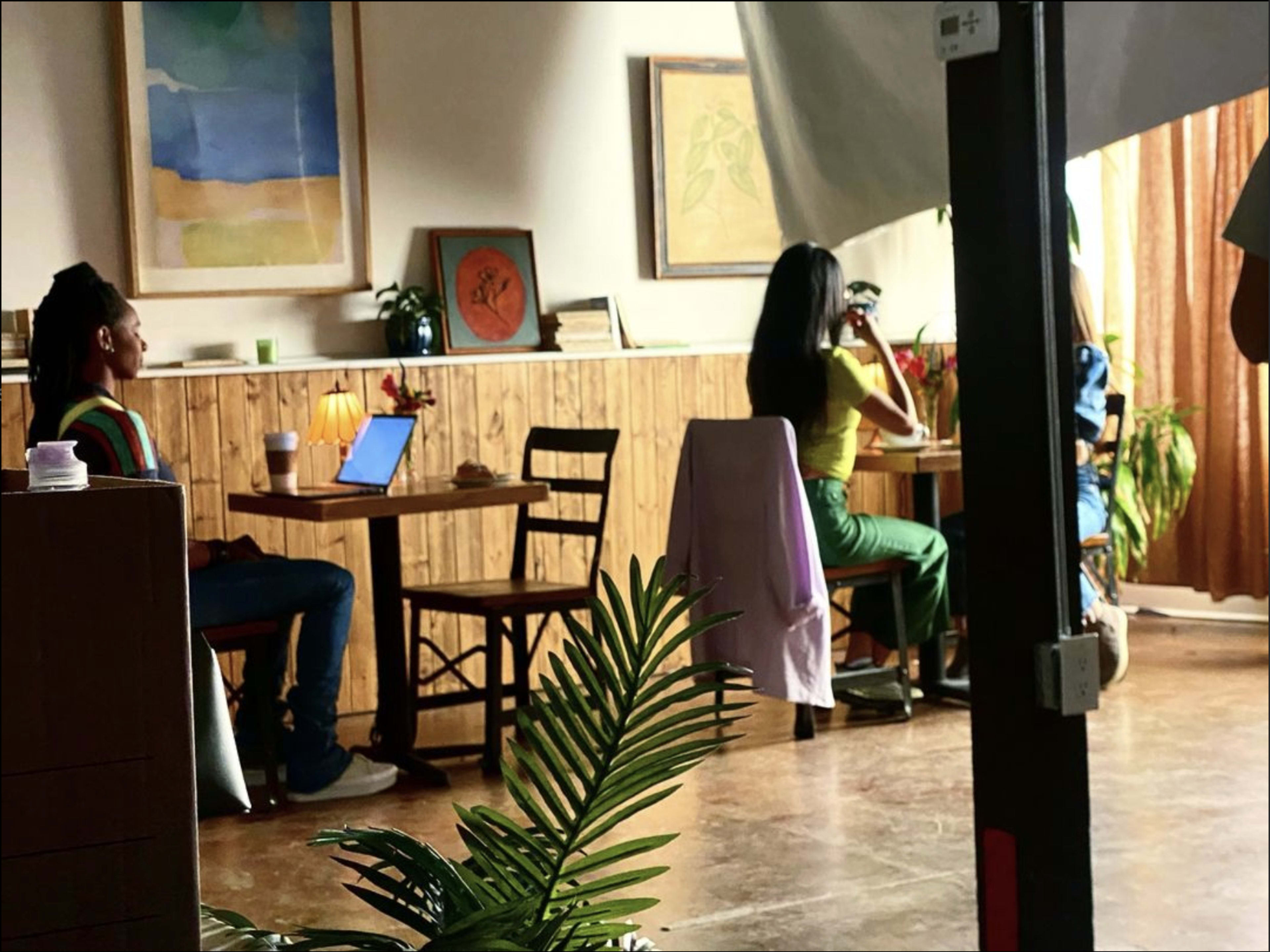 Two individuals are seated in a cozy café setting, one using a laptop at a table while the other sits nearby, sipping from a cup.