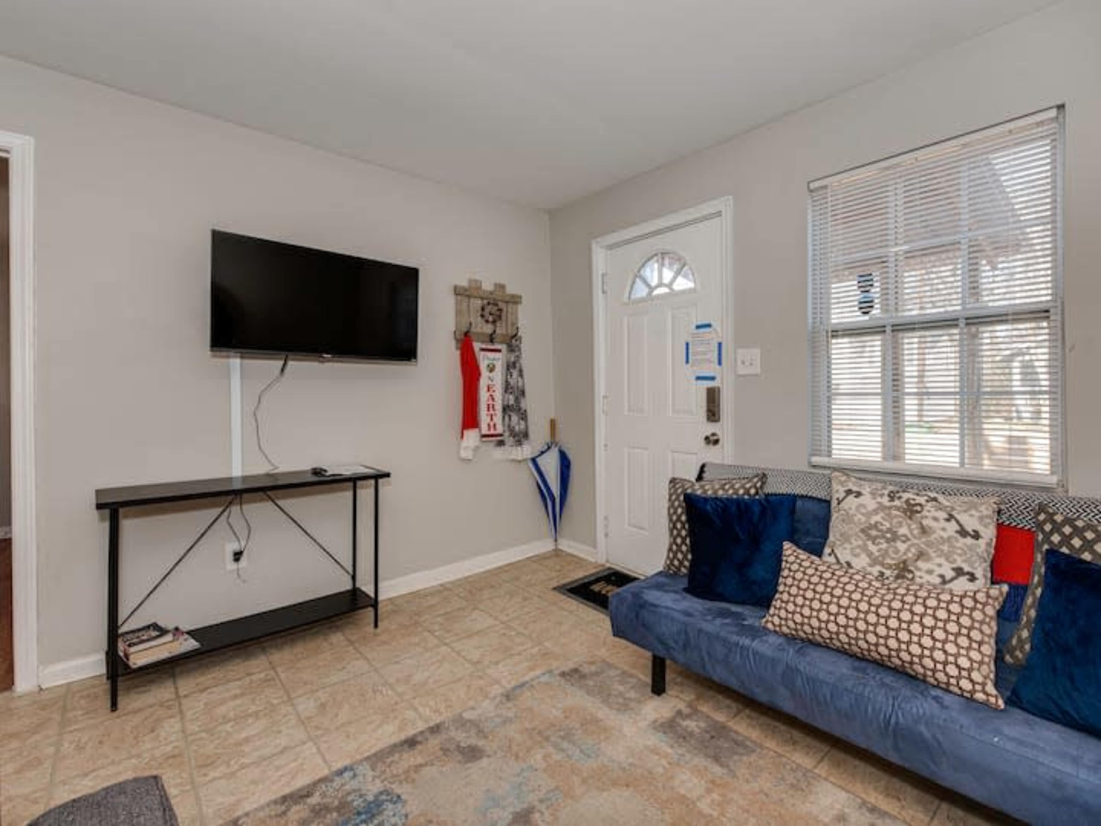 The room features a blue couch with decorative pillows, a wall-mounted television, and a door leading outside.