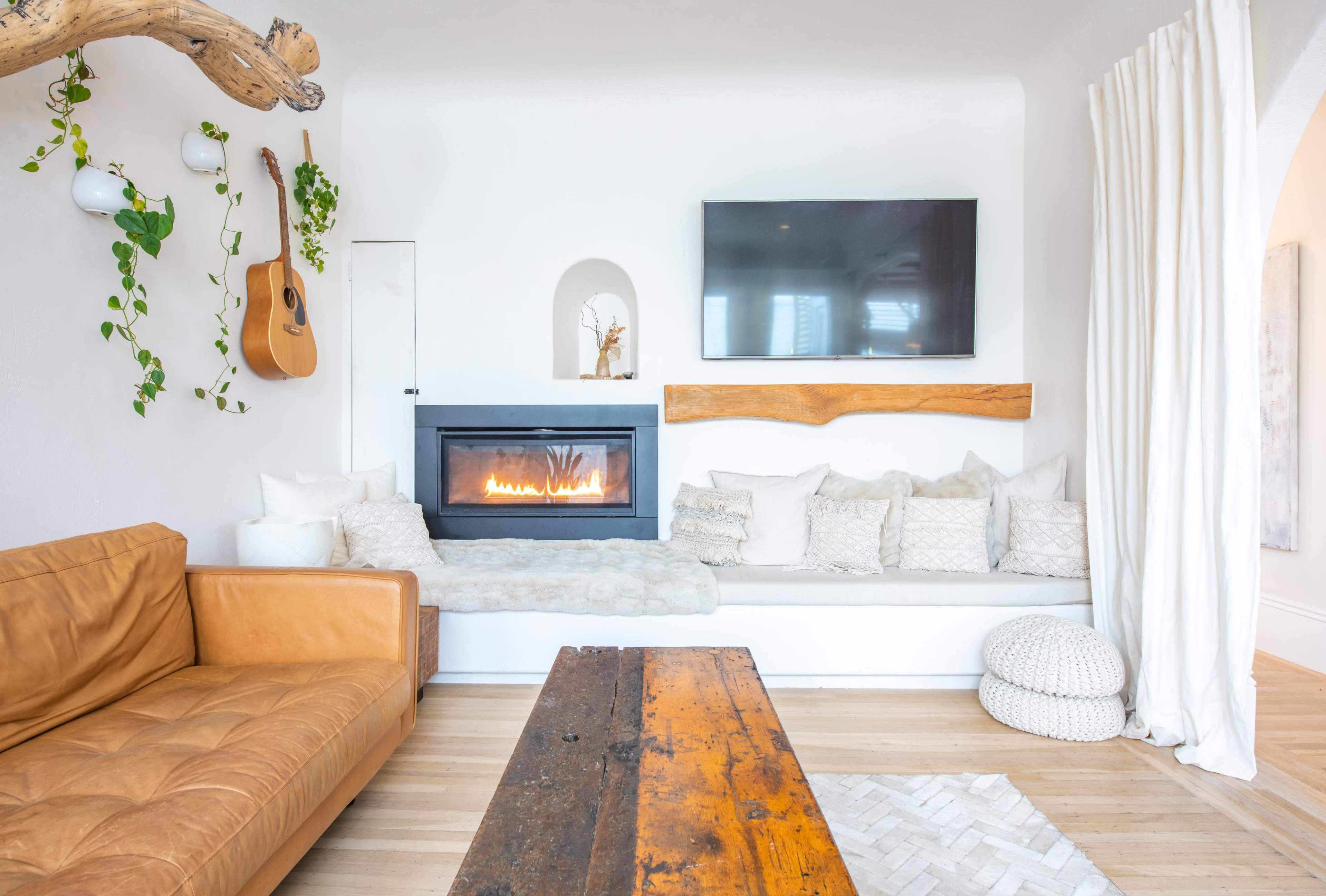 The image shows a modern living room featuring a fireplace with a wooden mantel, a light-colored sofa with numerous cushions, a wooden coffee table, and decorative plants on the walls.