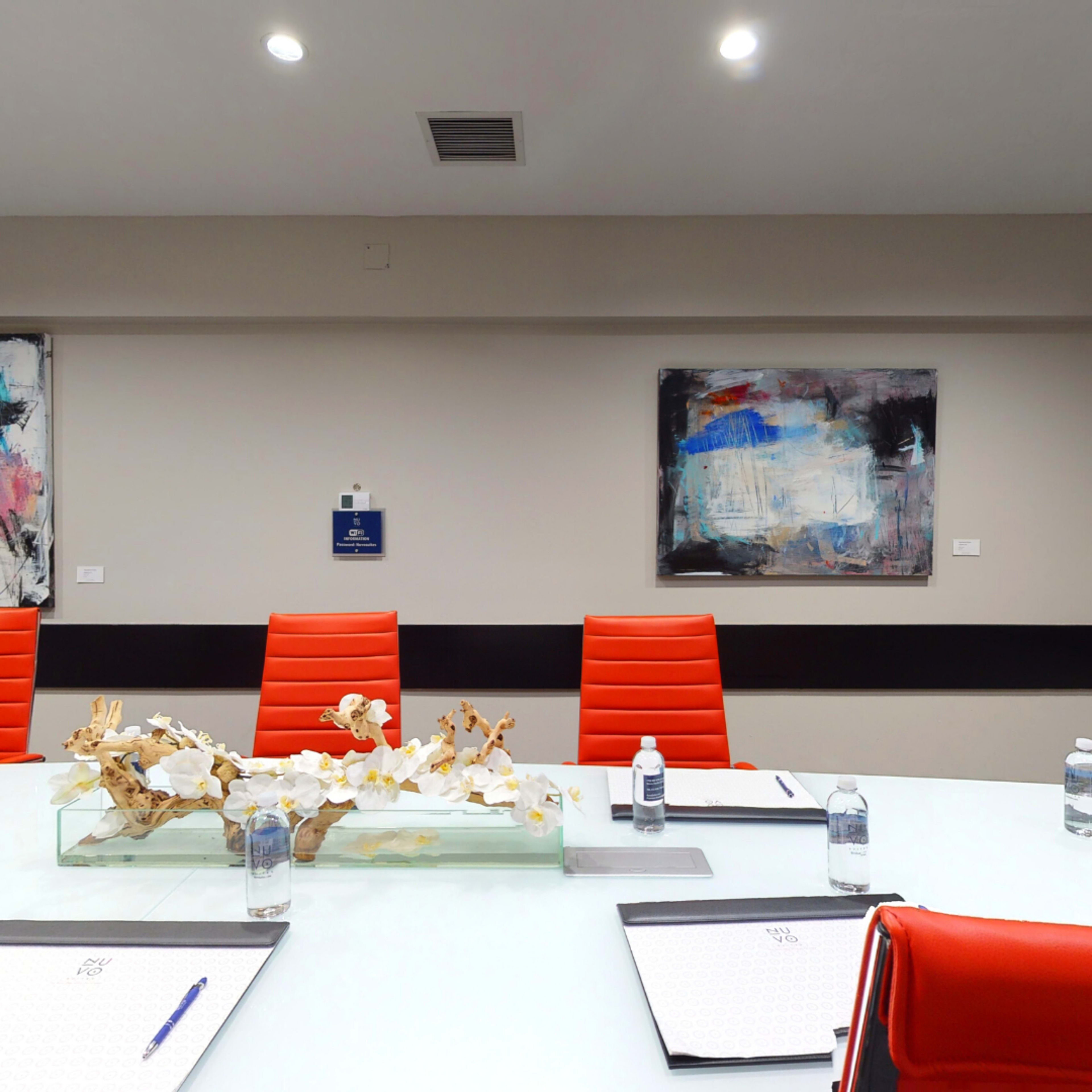 A modern conference room features a glass table with a decorative centerpiece, eight red chairs, and water bottles on white notepads.
