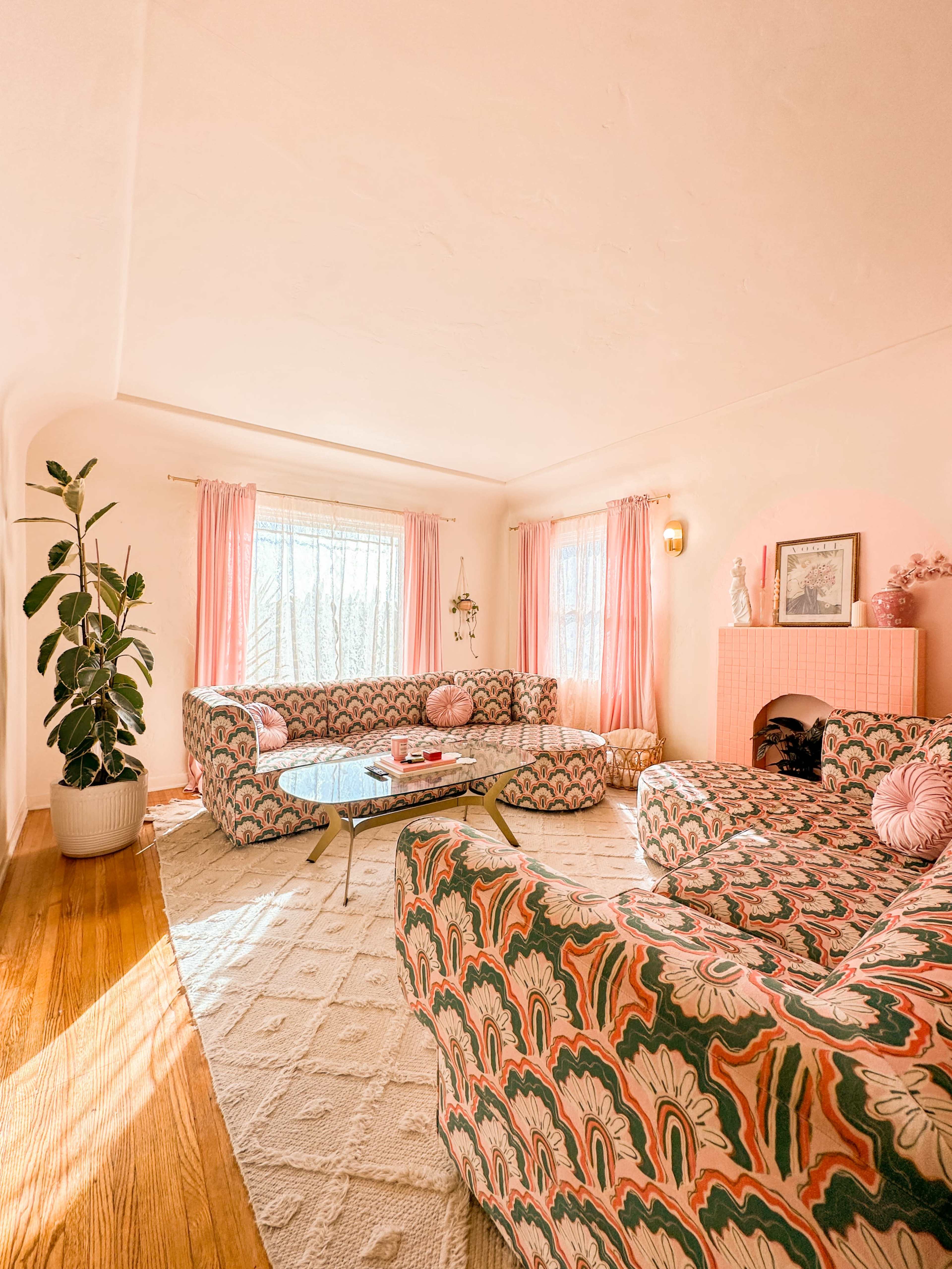 The living room features two large patterned sofas, a glass coffee table, and a potted plant, all set against pale pink walls and large windows letting in natural light.