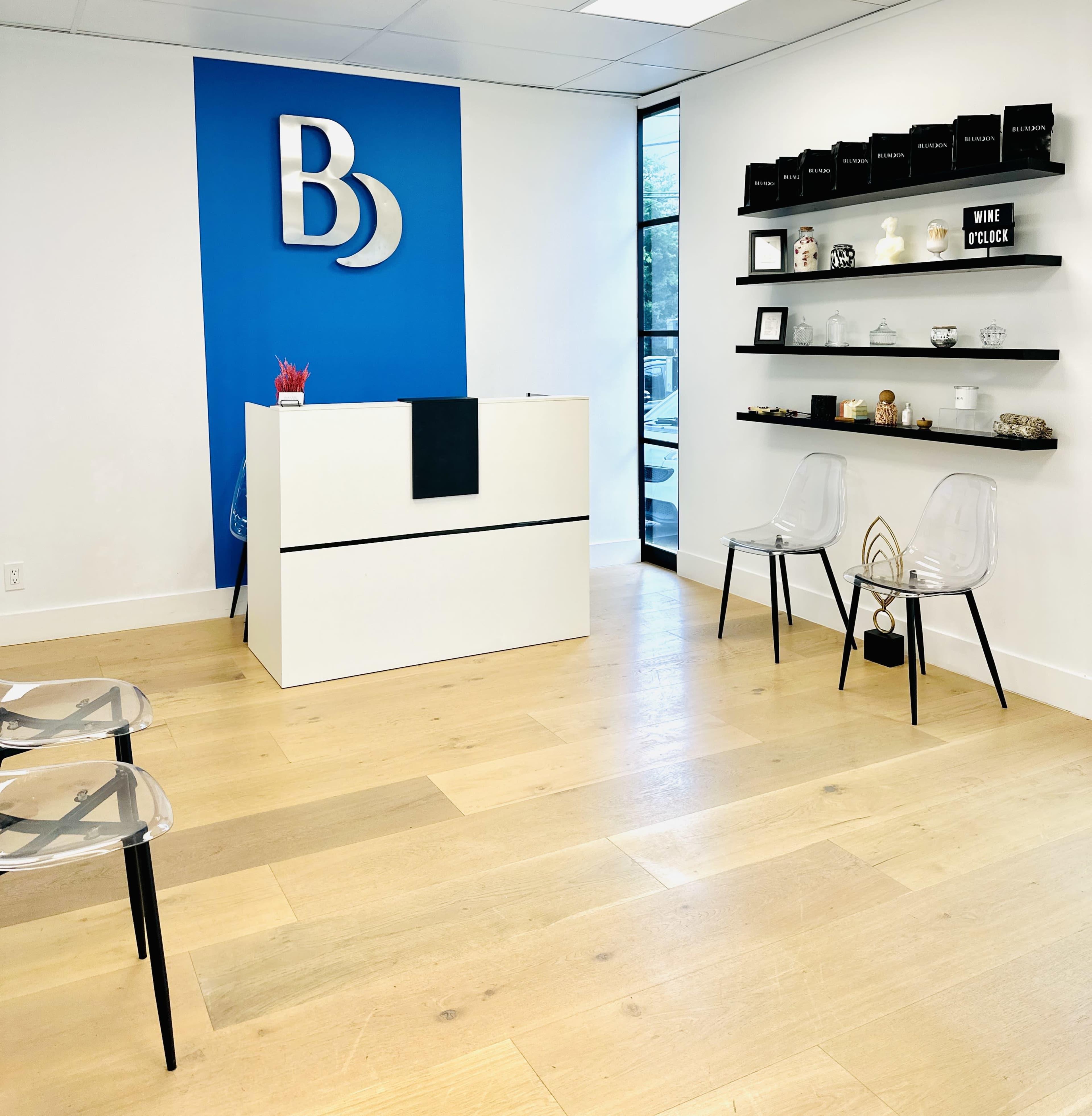 The image shows a modern reception area with a white desk, a blue wall featuring a logo, and a display shelf holding various items, alongside two transparent chairs.