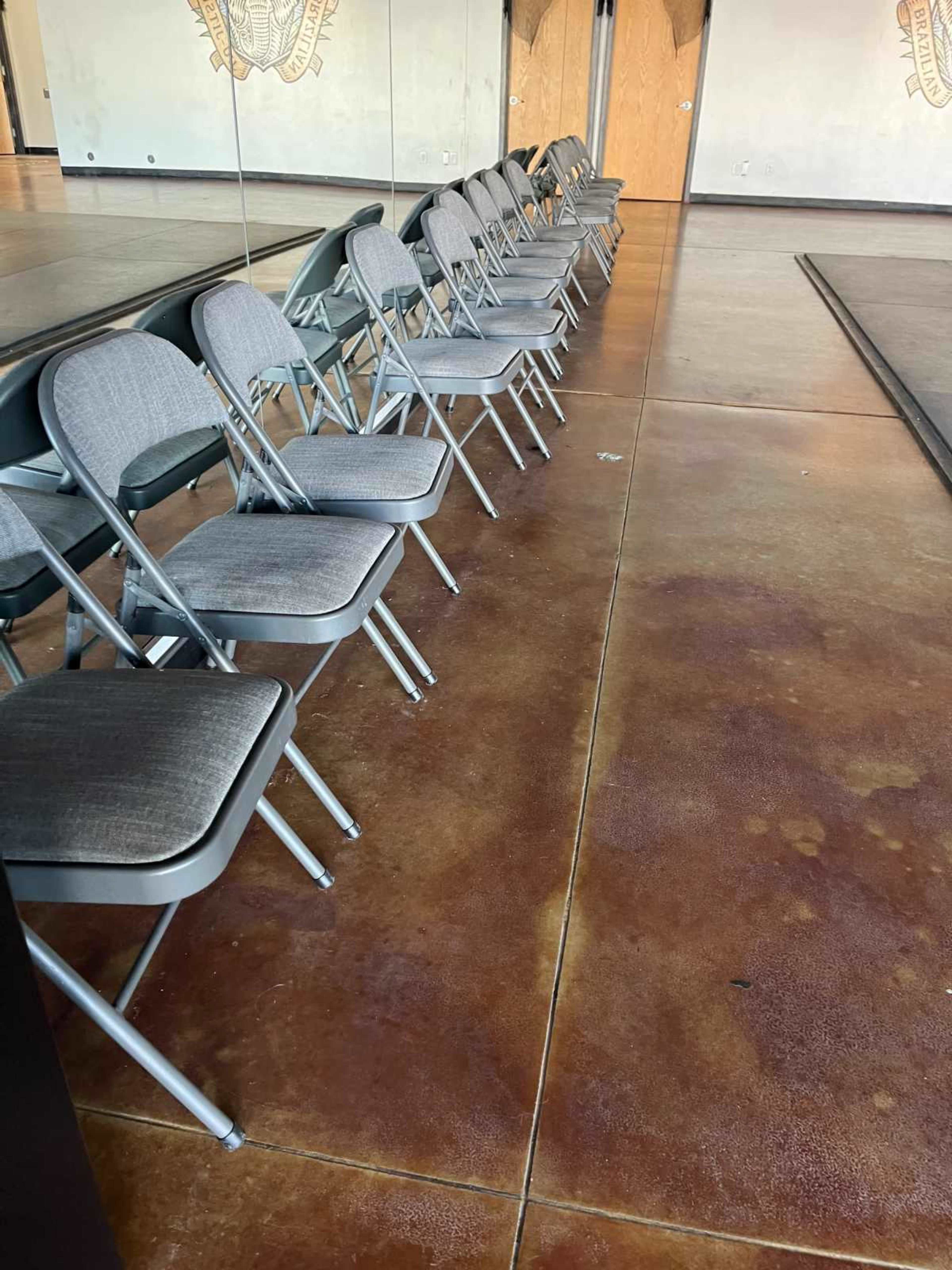 A row of gray folding chairs is aligned against a wall in a studio space with polished concrete flooring.