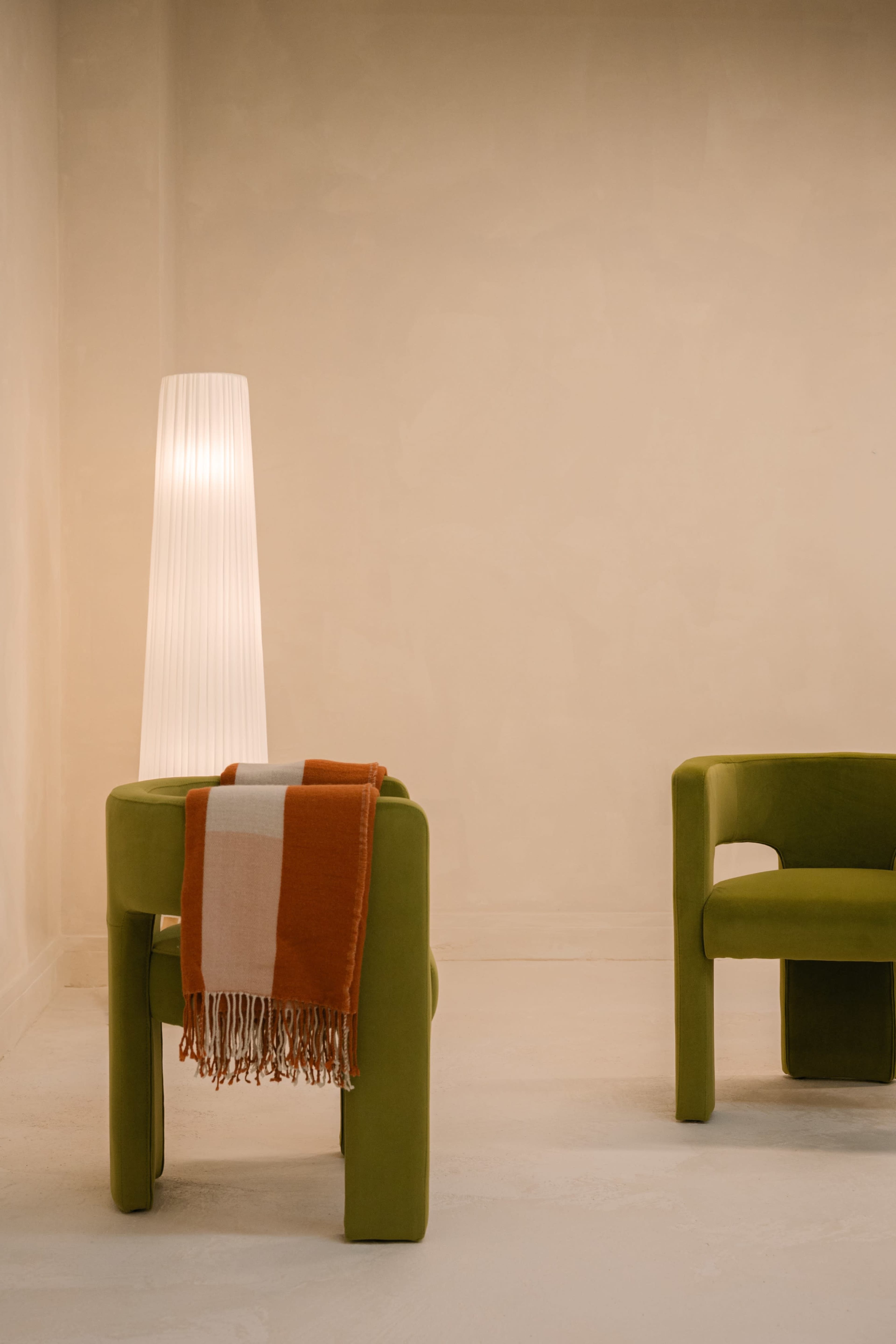 A minimalistic interior features two green chairs, one draped with an orange and white blanket, beside a tall, textured lamp against a beige wall.