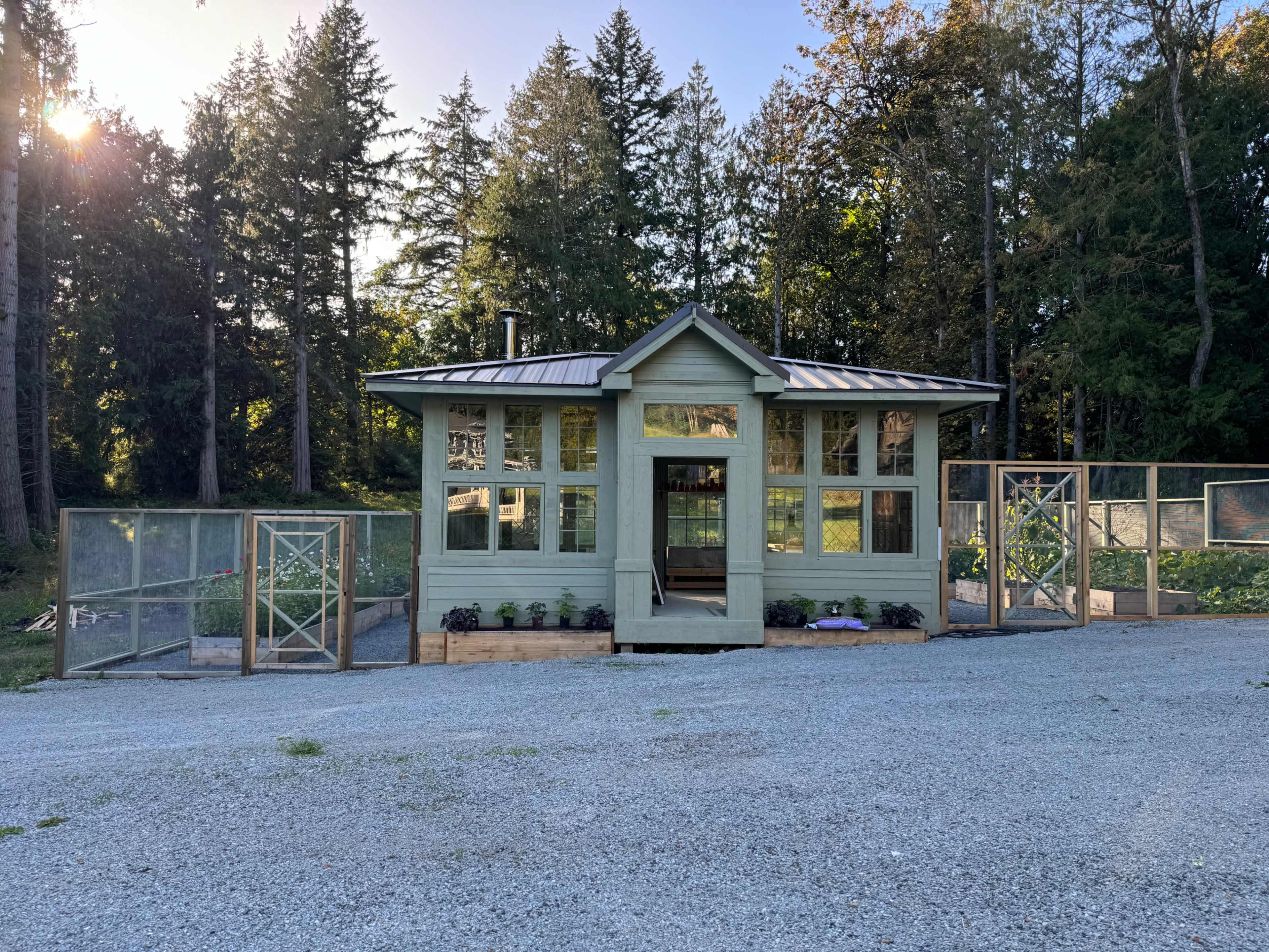 A small green building with large windows is situated in a gravel area, surrounded by wooden fencing and trees.