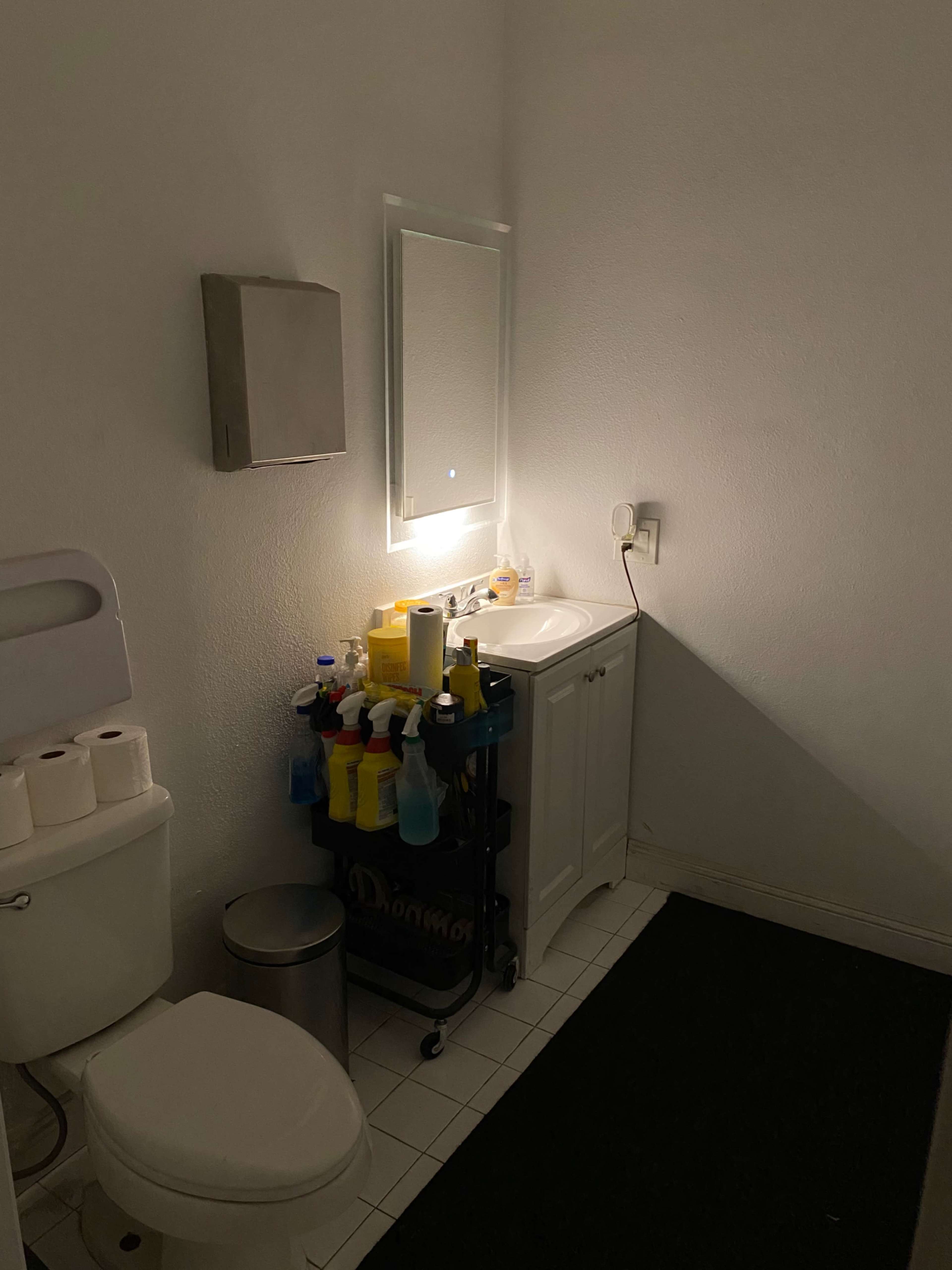 A dimly lit bathroom features a toilet, a small sink with a mirror, and a cart filled with cleaning supplies.