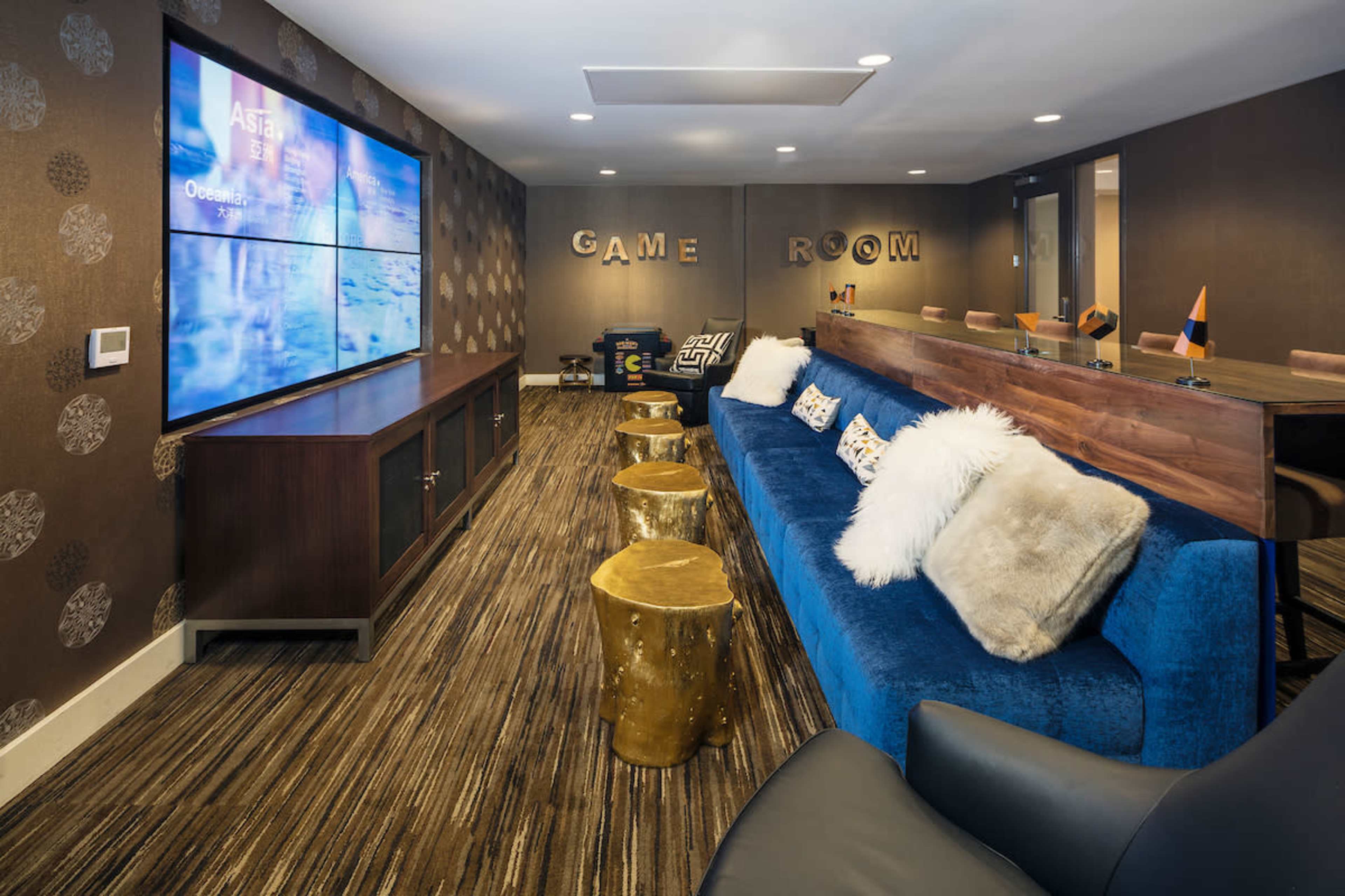 A game room features a blue sofa, gold accent stools, a long wooden table, and large screens displaying content on the walls.