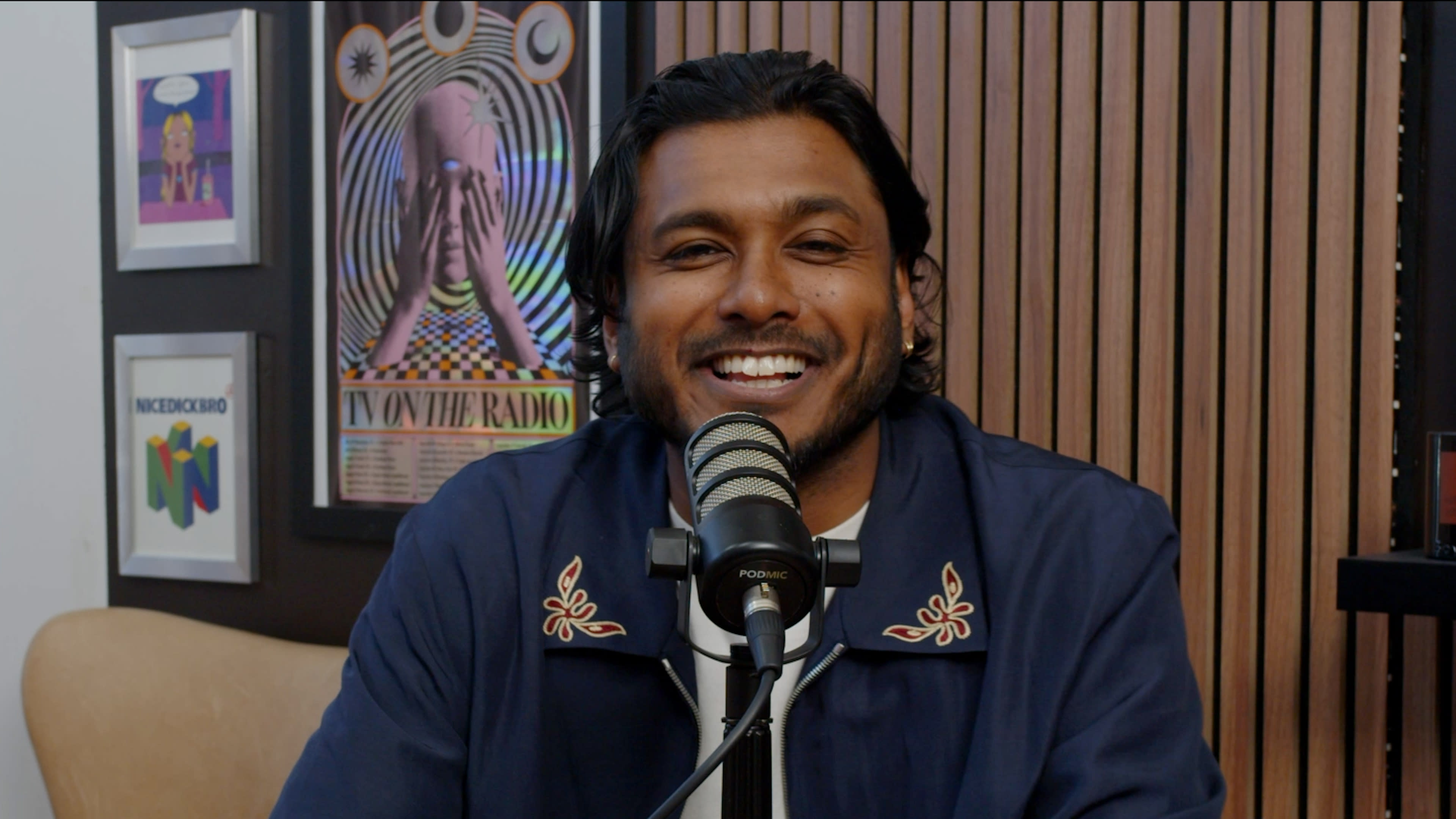 A man with dark hair and a blue jacket smiles while sitting in front of a microphone, with colorful artwork on the wall behind him.