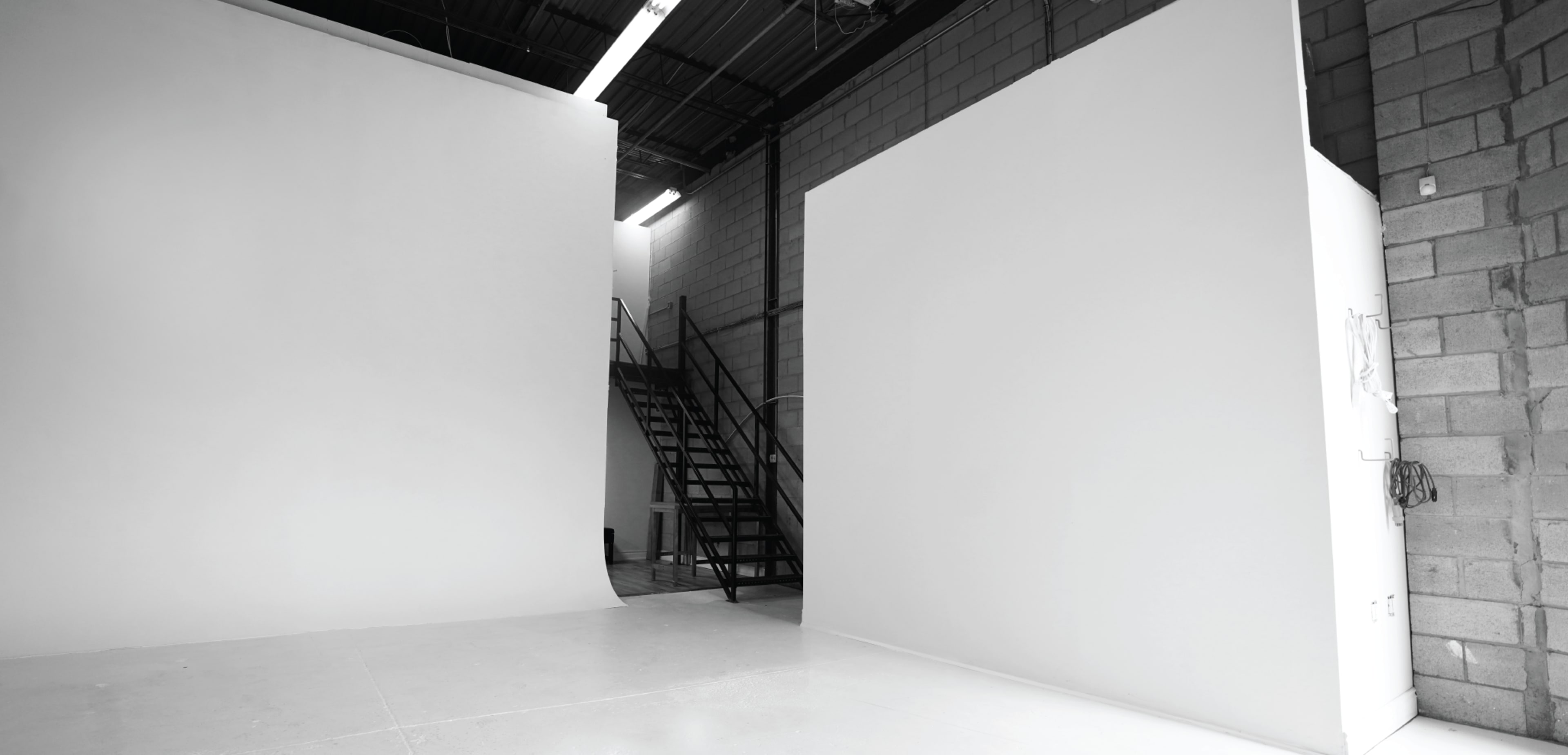 A spacious, empty photo studio with two white walls and a black staircase in the background.