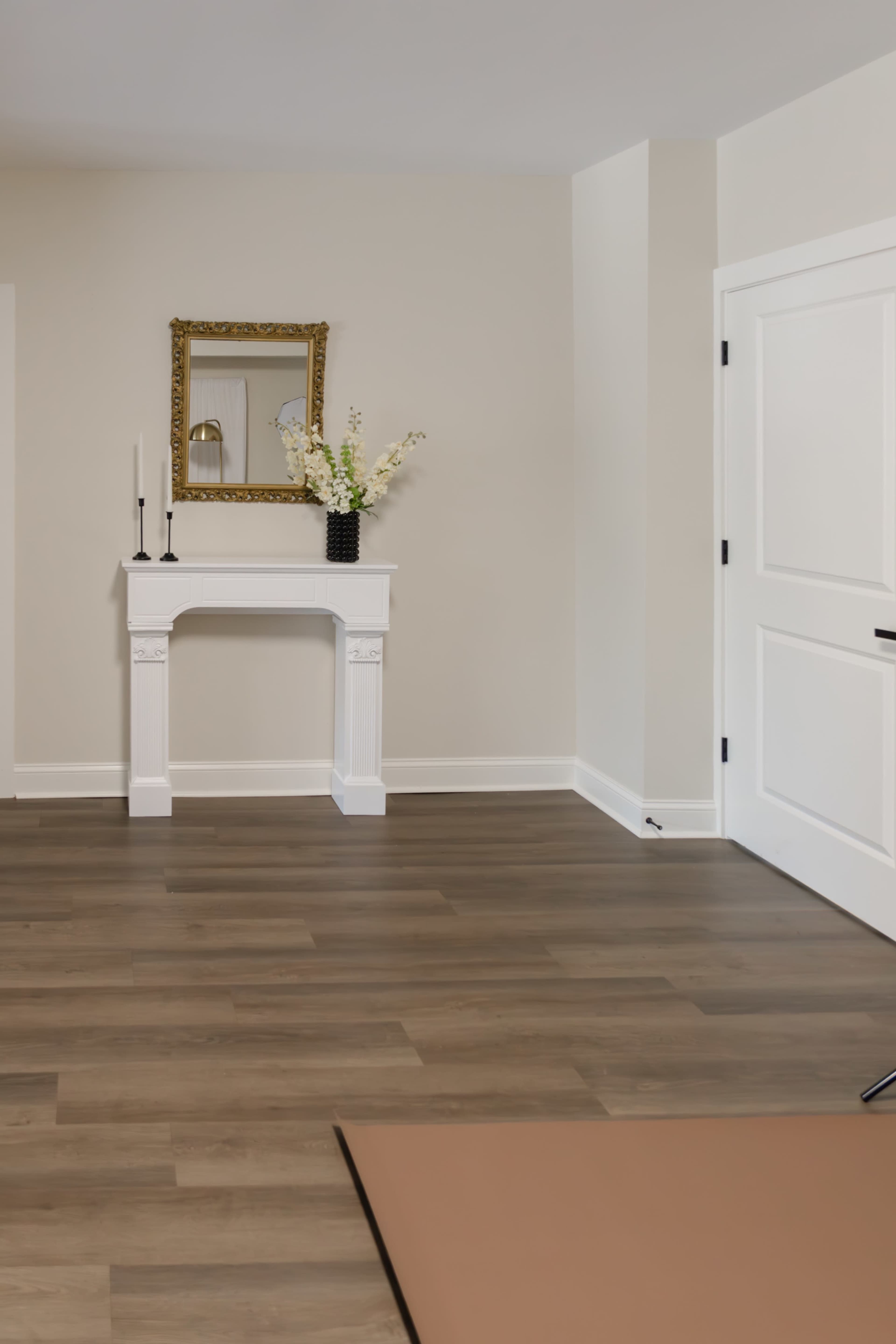 The image depicts a minimalist room with a wooden floor, a decorative fireplace with a mirror above it, and a door on the right side.
