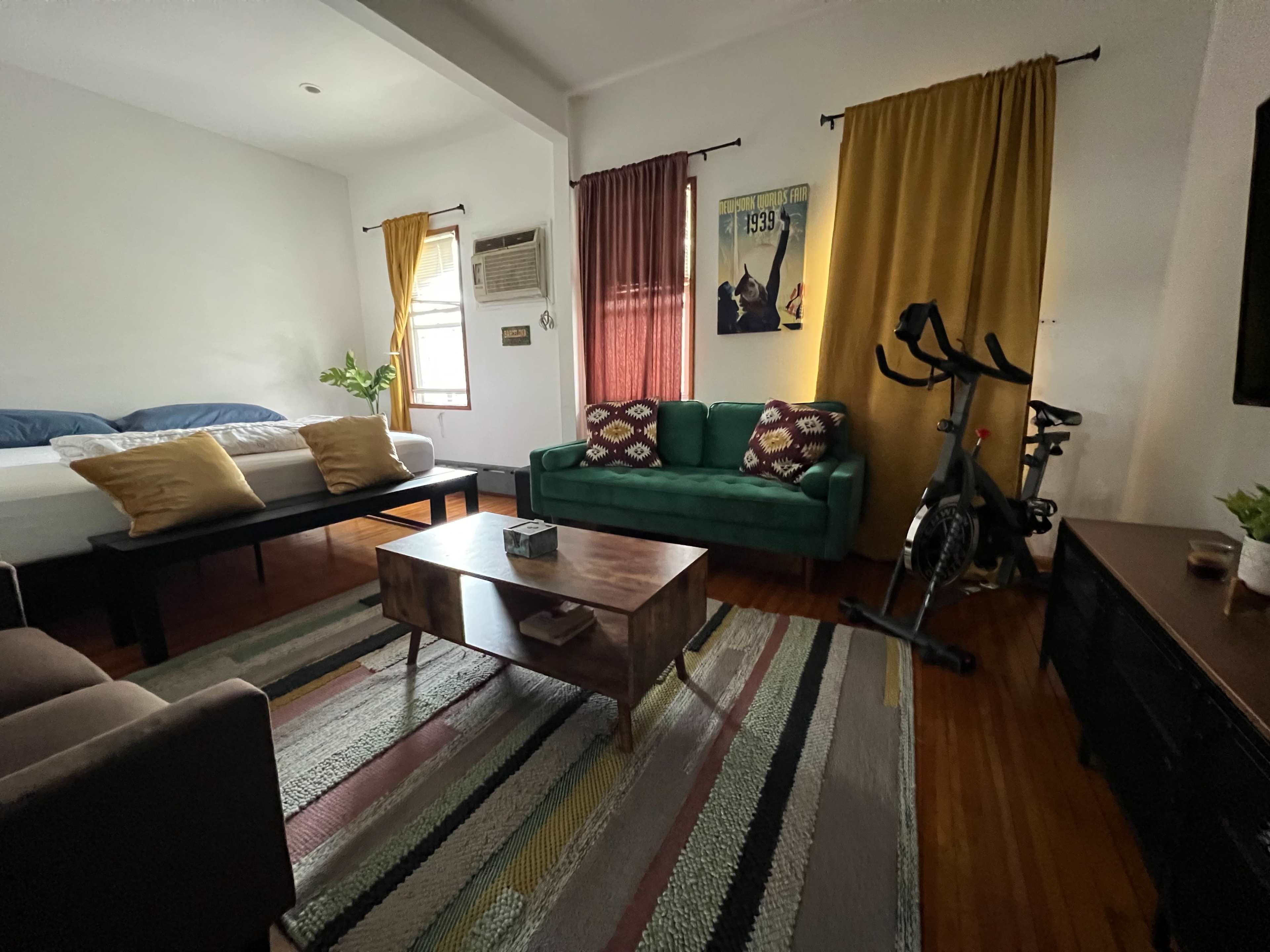 The image shows a living room with two beds in the background, a green sofa, a coffee table, an exercise bike, and colorful curtains.