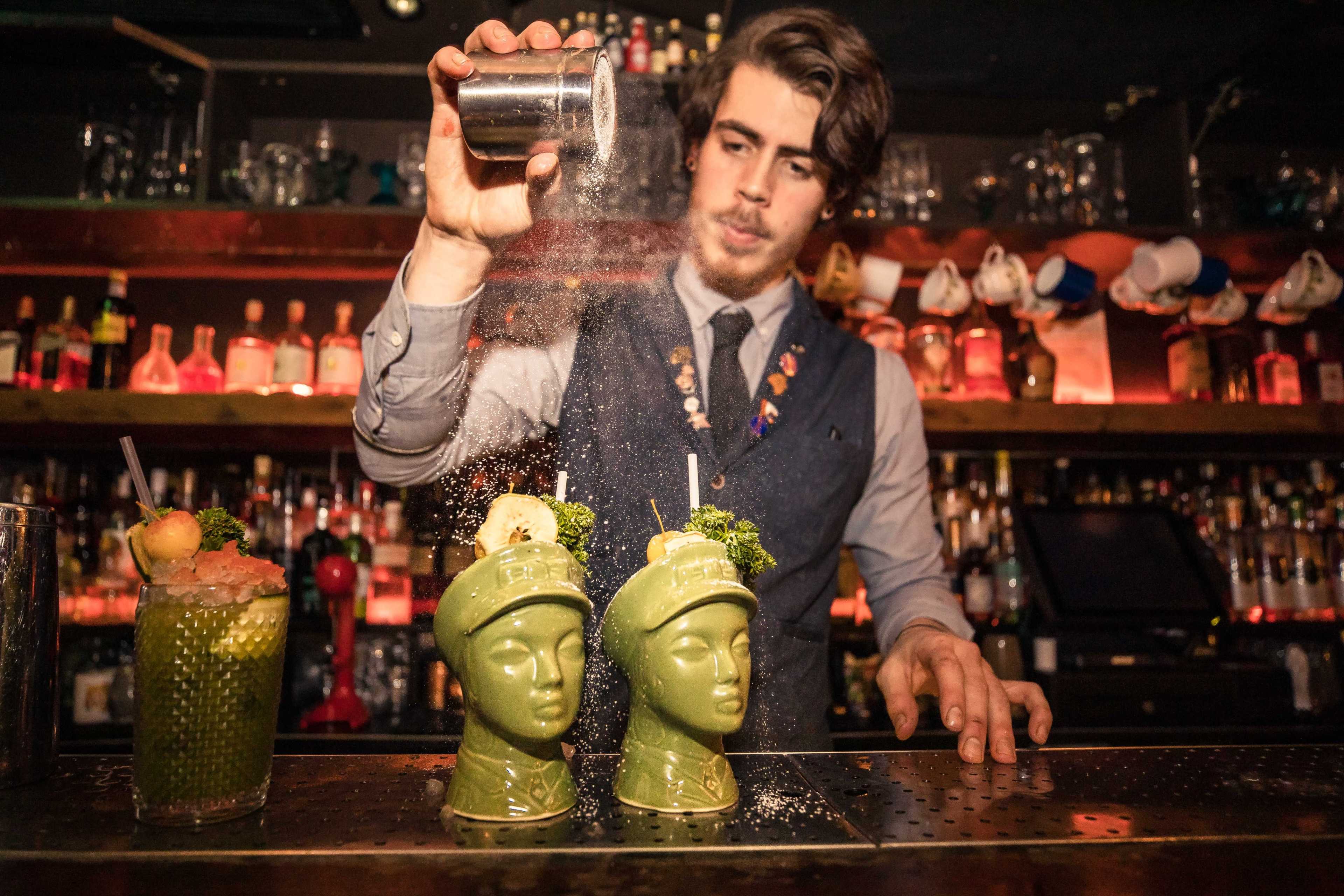 A bartender sprinkles sugar over two green cocktail mugs shaped like faces while mixing drinks at a bar.