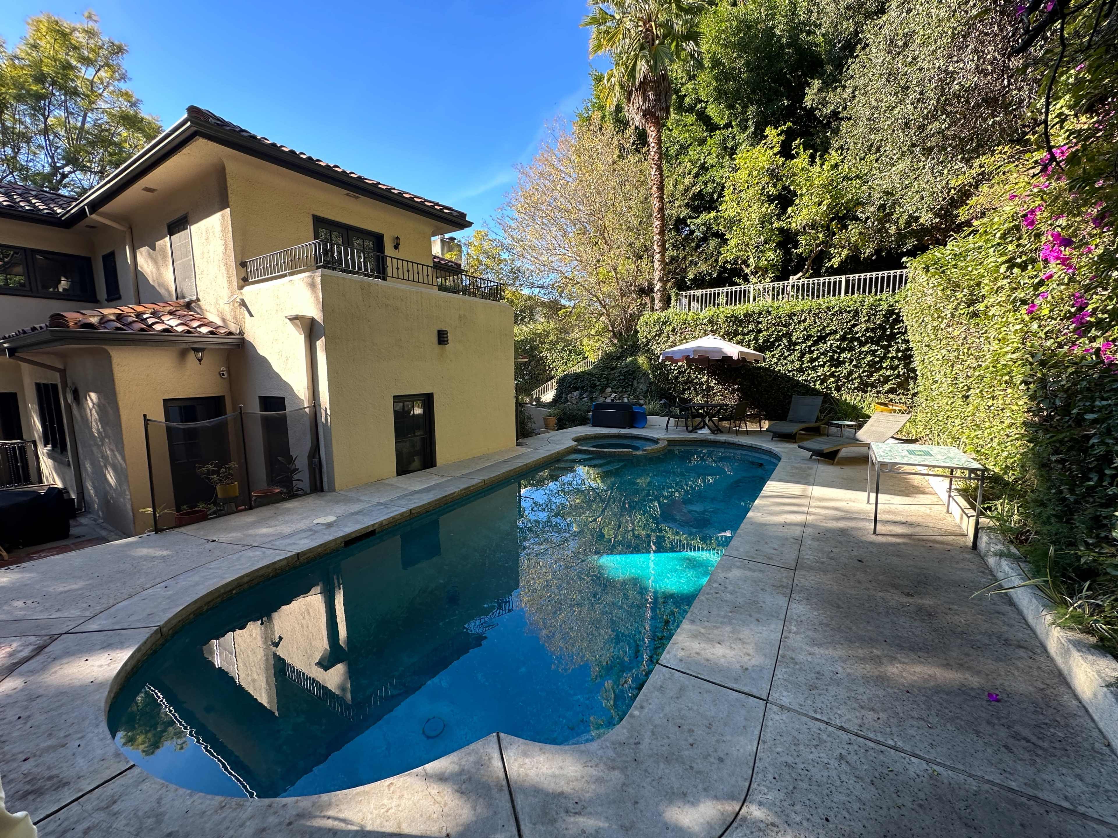 A backyard poolside area features a curved swimming pool surrounded by concrete, lounge chairs, and a two-story house amidst lush greenery.