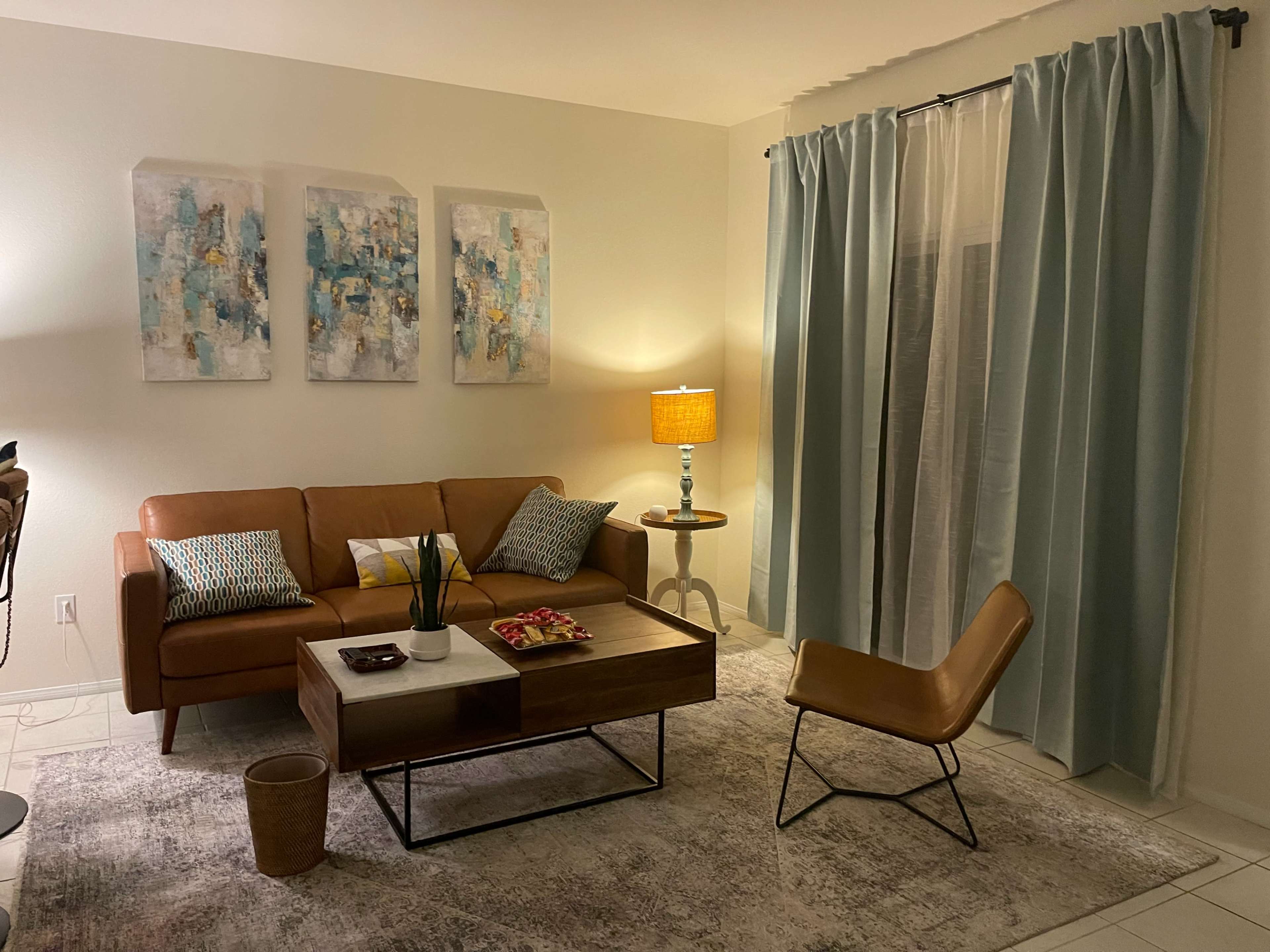 A living room features a brown sofa with colorful cushions, a wooden coffee table, and a light-colored rug, accompanied by a floor lamp and blue curtains.