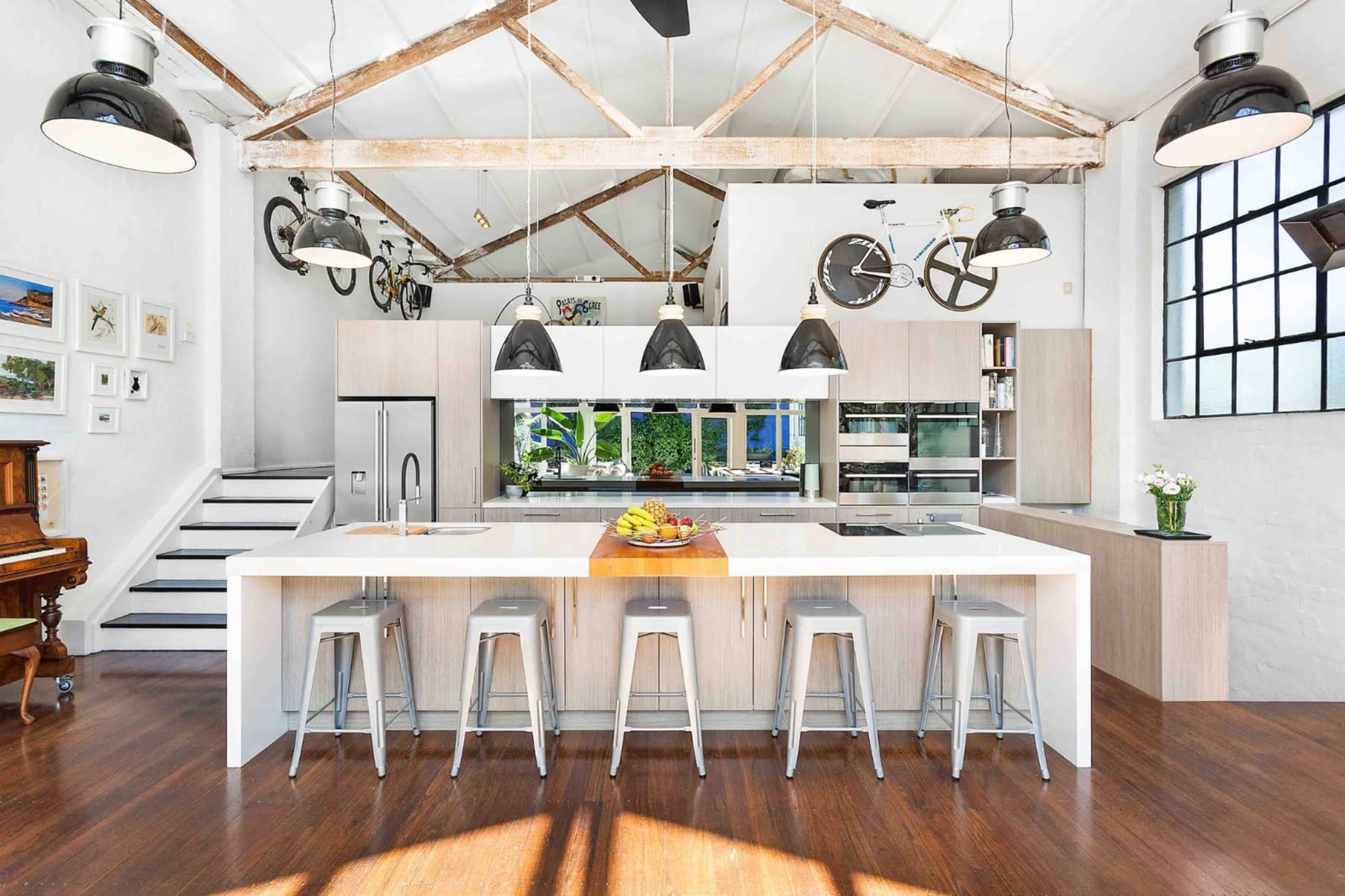 The image shows a spacious kitchen with a large central island, modern appliances, and a high ceiling featuring exposed beams and pendant lighting.