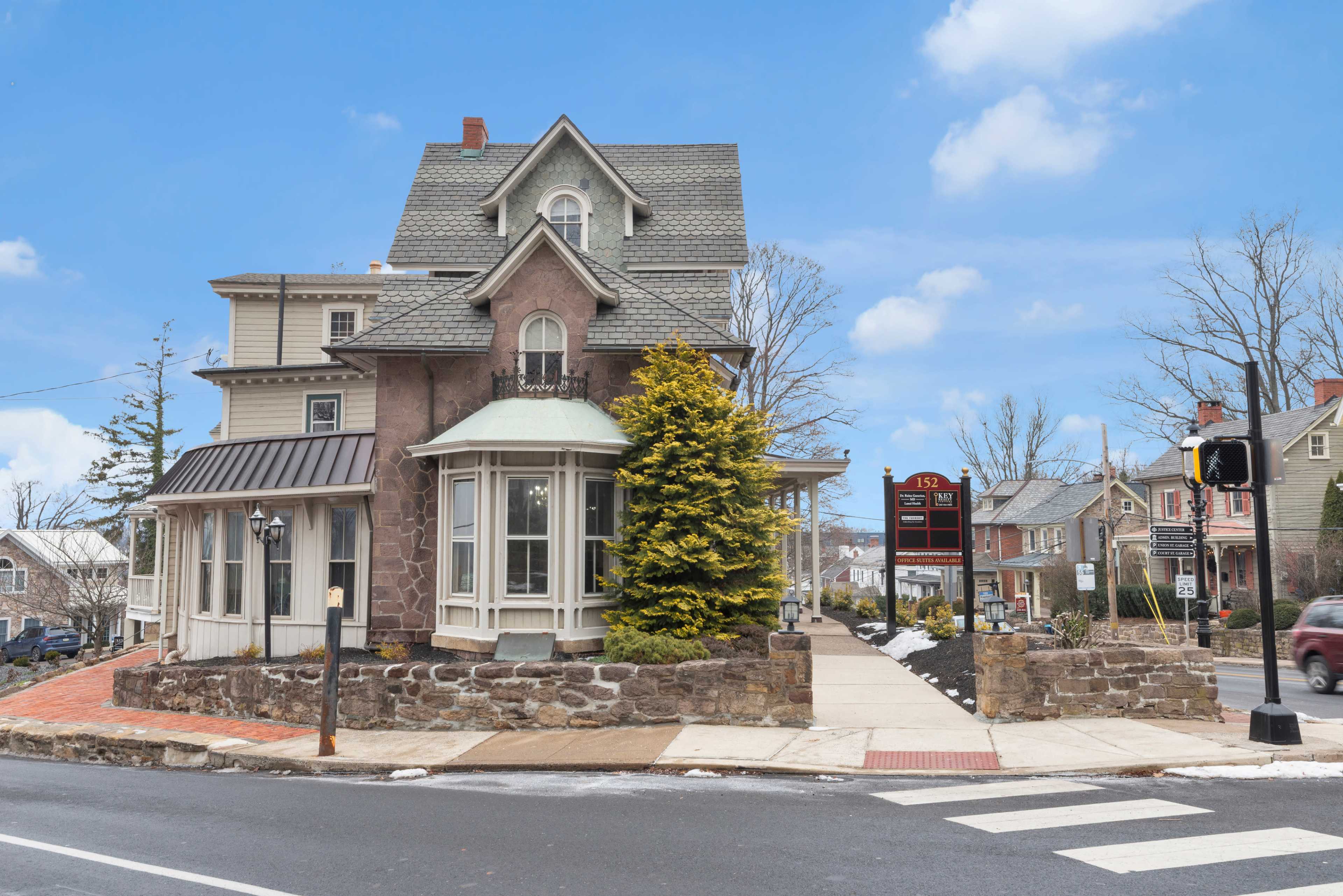A two-story house with a distinctive turret and bay window sits at an intersection, surrounded by trees and street signage.