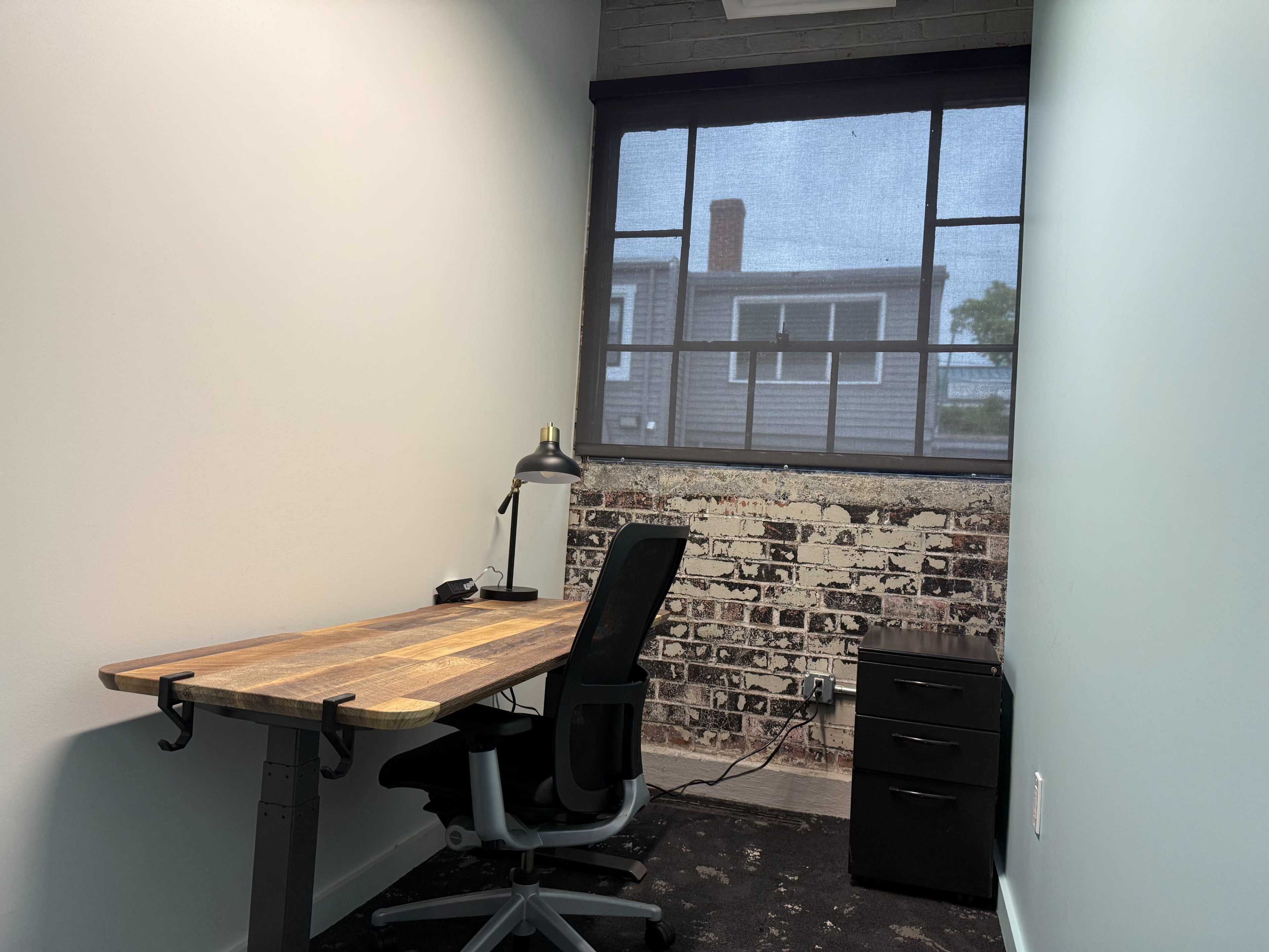 The image shows a small office space with a wooden desk, an ergonomic chair, and a dark filing cabinet against a wall featuring a brick design, with a window casting natural light into the room.