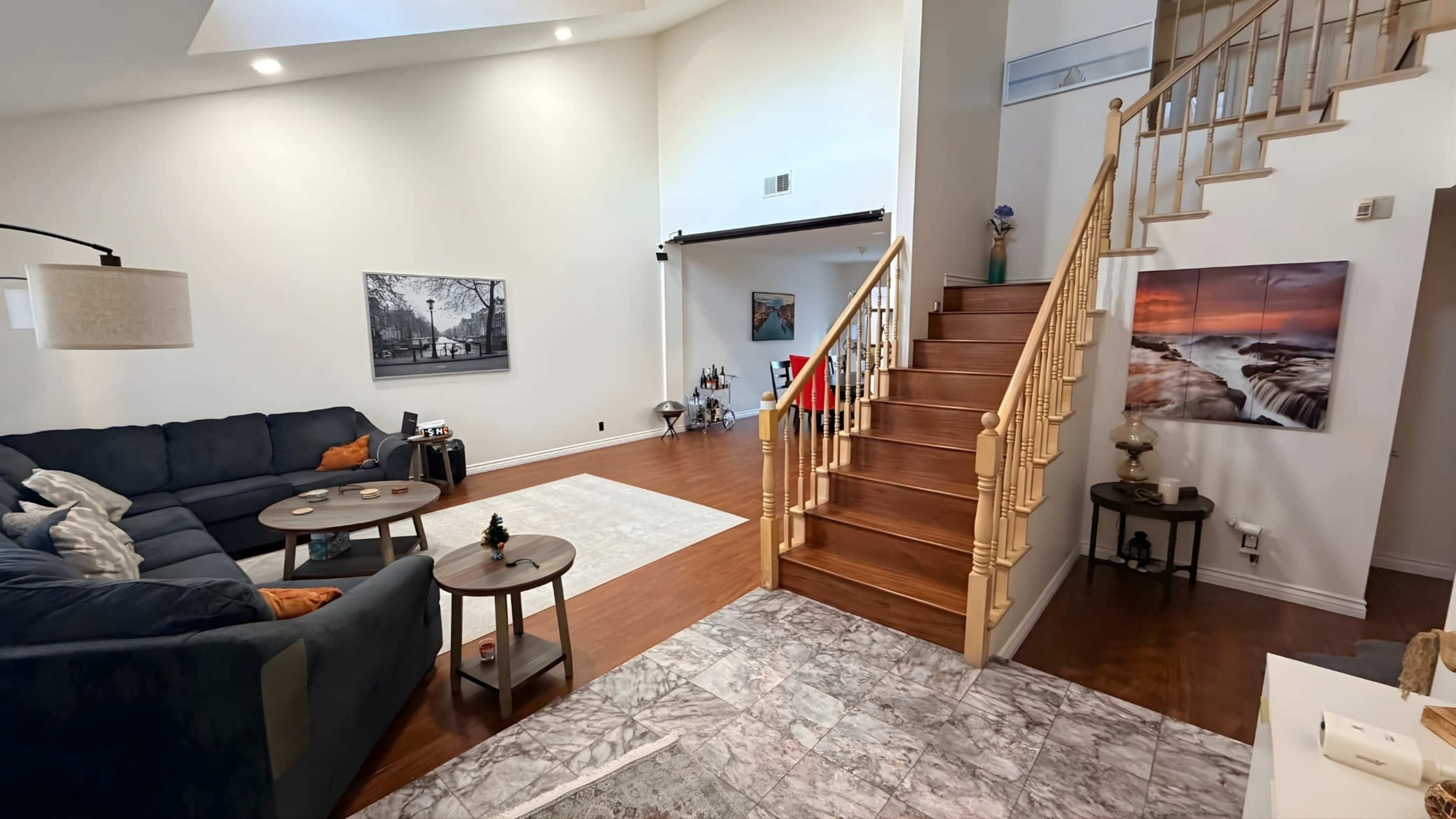 The image shows a spacious living area with a dark sofa, a set of small tables, and a staircase leading to an upper level, decorated with various artworks and a rug on the floor.
