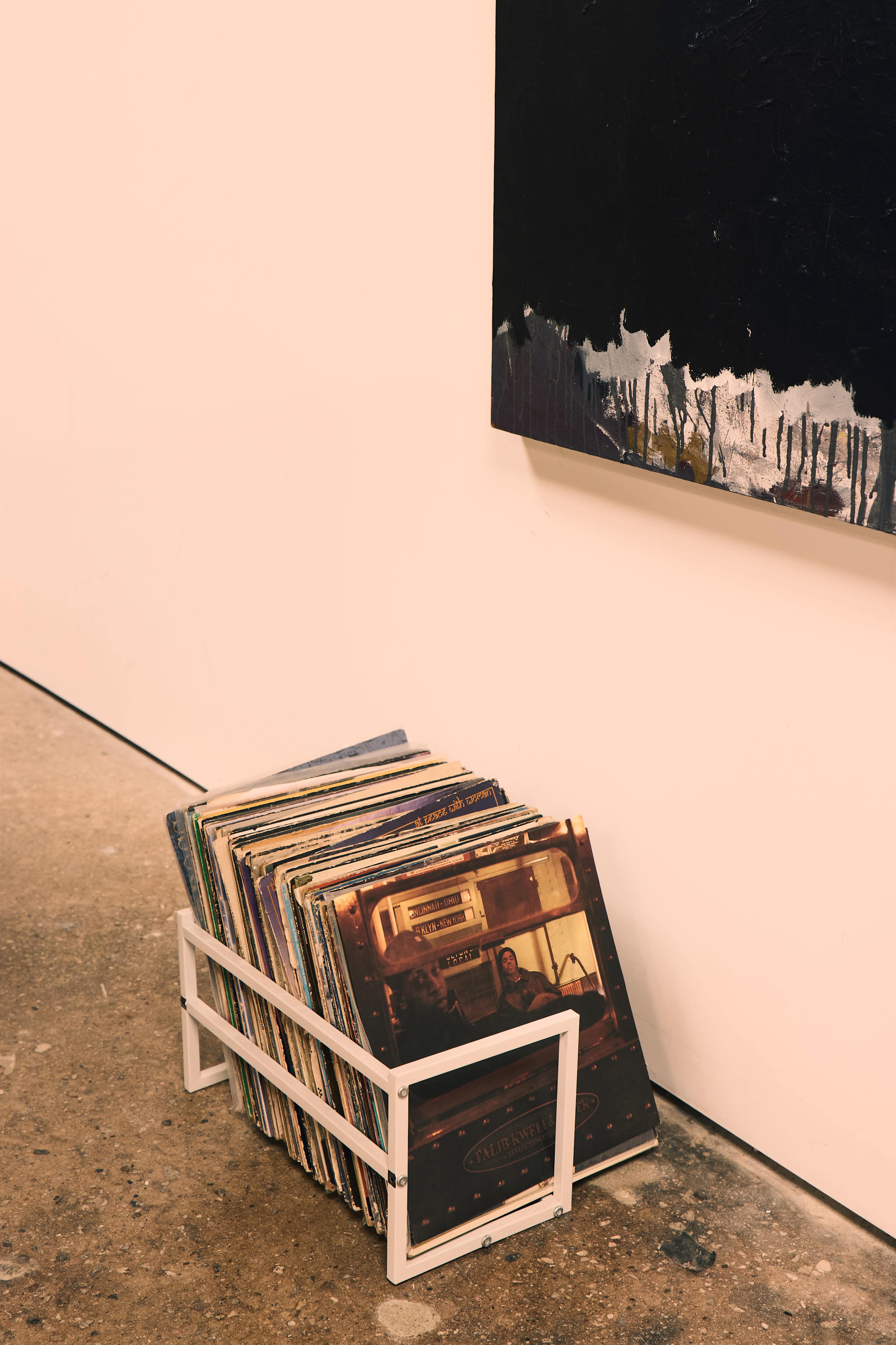 A metal record holder containing various vinyl records is placed against a wall next to a large abstract painting.