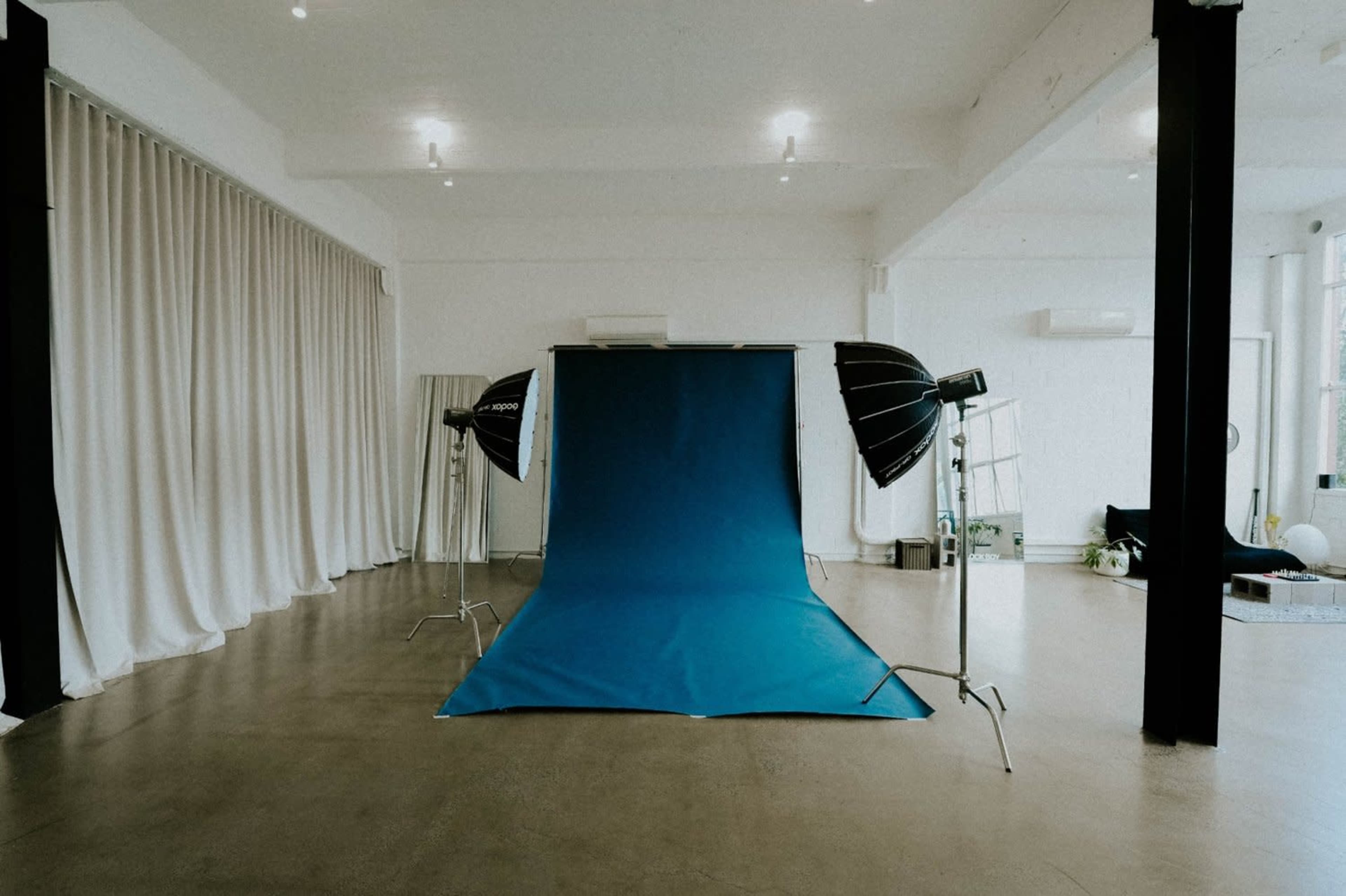 The image shows a photography studio with a blue backdrop, large light modifiers, and white curtains in a minimalistic space.