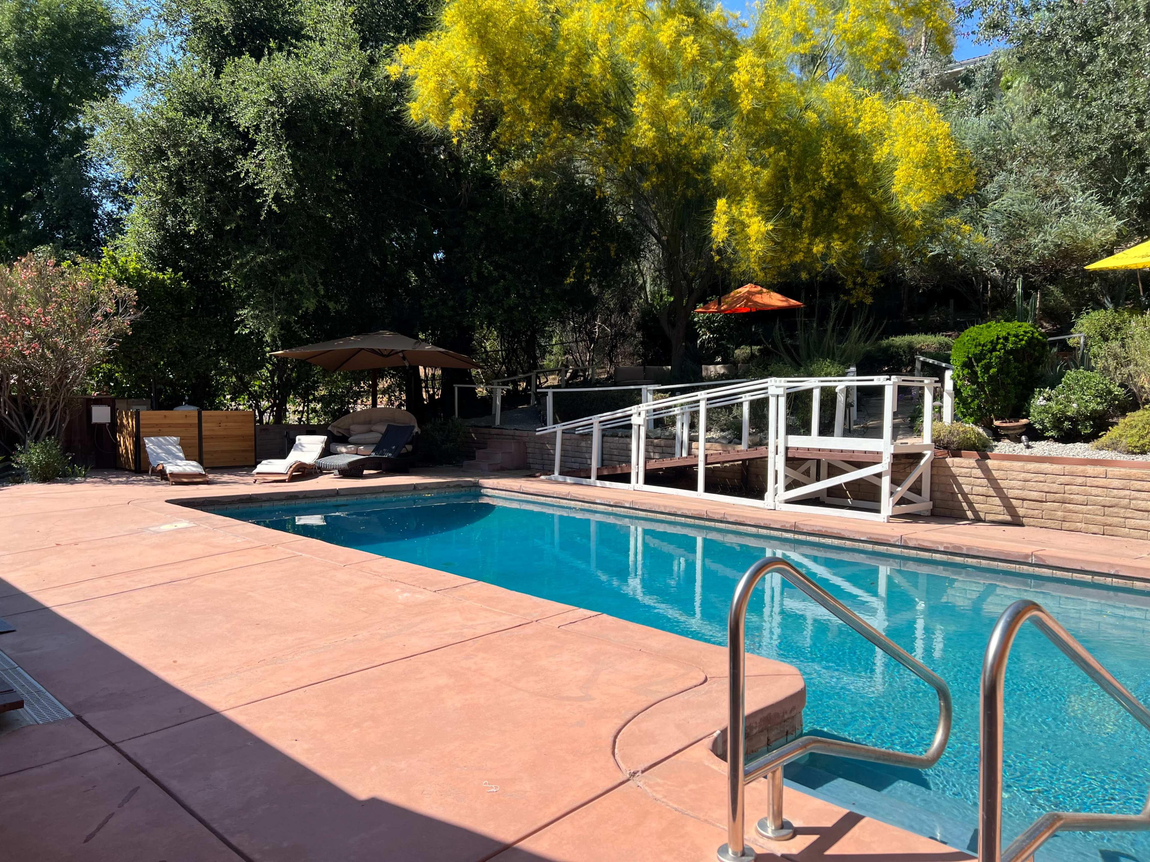 A swimming pool surrounded by lounge chairs, a shaded cabana, and landscaped greenery under a clear blue sky.