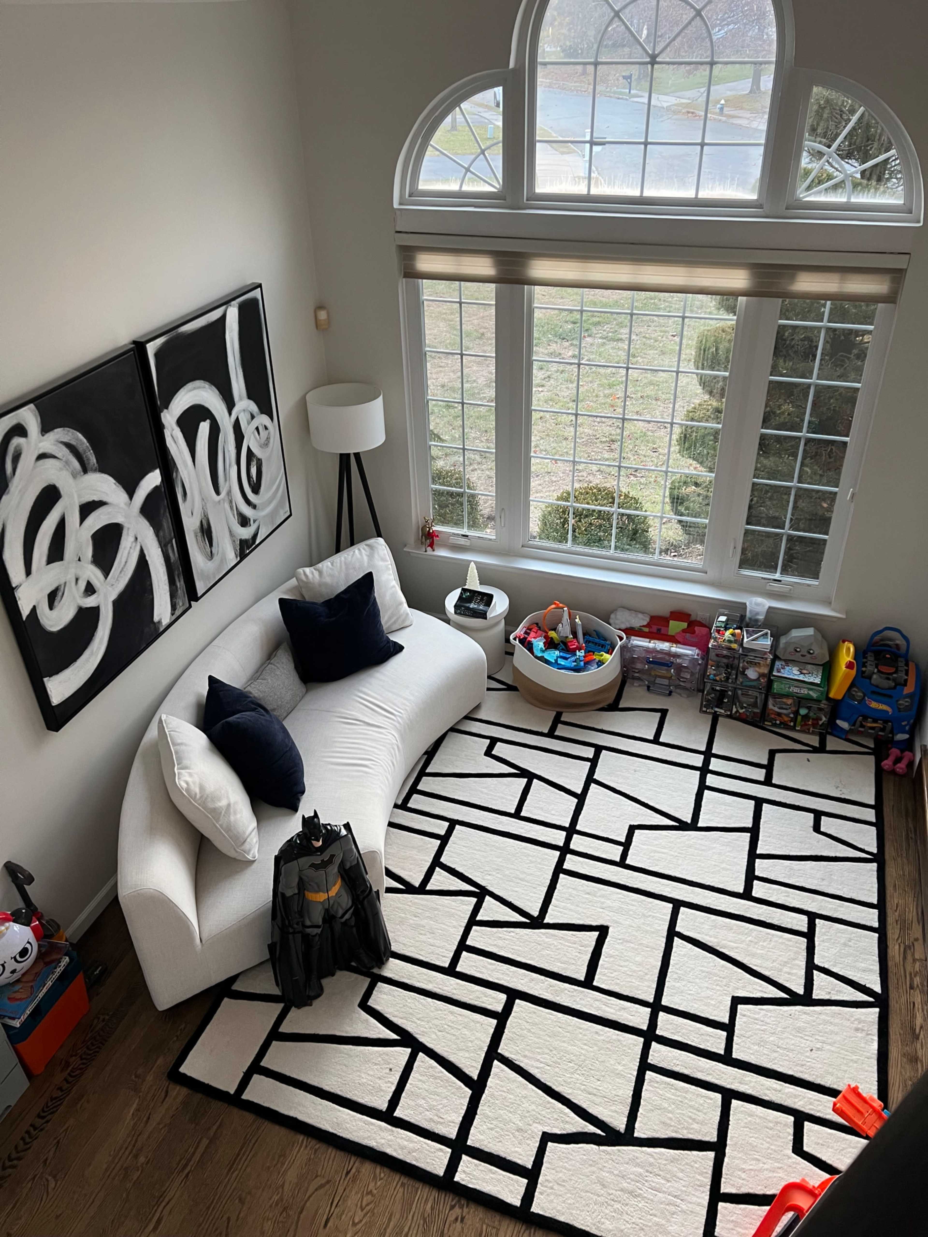 The image shows a bright room with a large window, a white sofa with navy blue pillows, a geometric rug, and various toys organized on shelves.