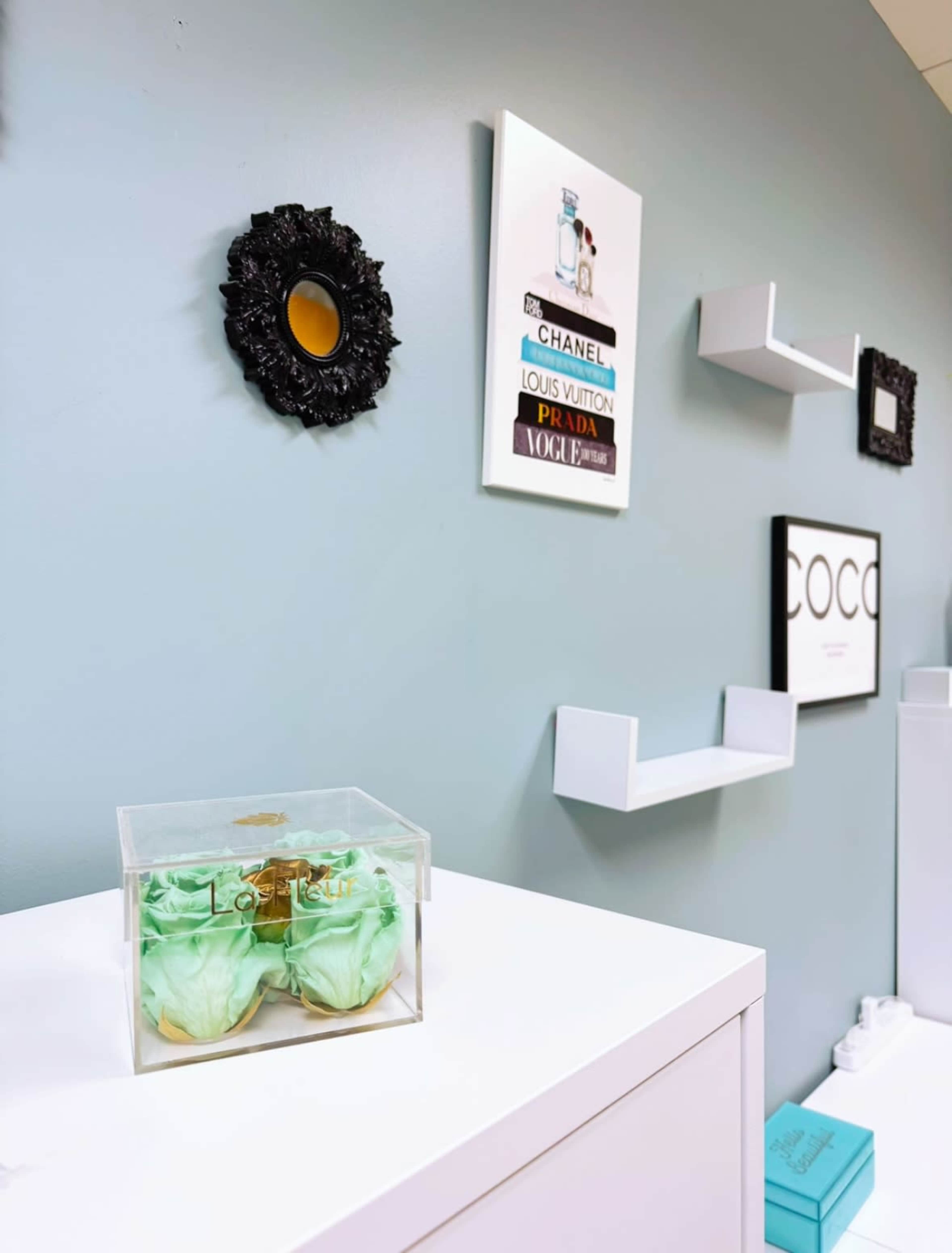 The image shows a light blue wall adorned with decorative shelves, framed art, and a clear display case containing a pair of green decorative sunglasses.