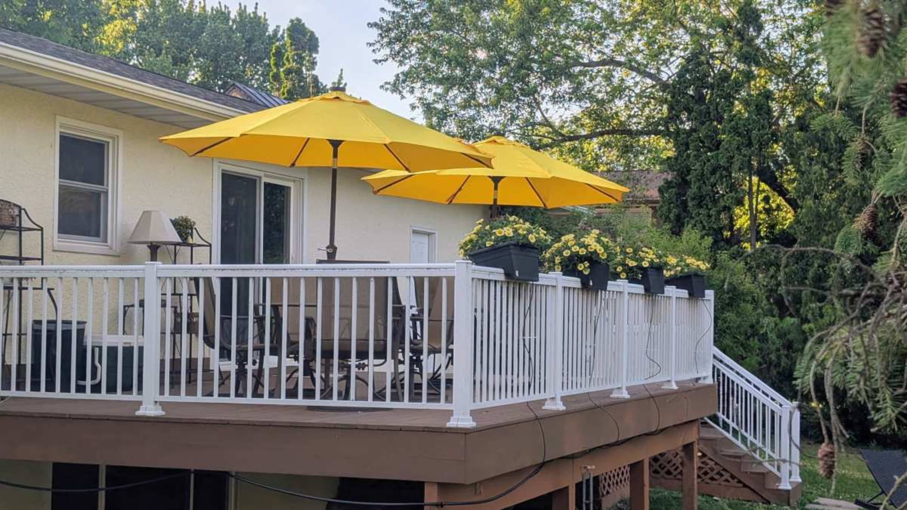 A wooden deck features yellow umbrellas over a seating arrangement and flower pots with yellow blooms along the railing.