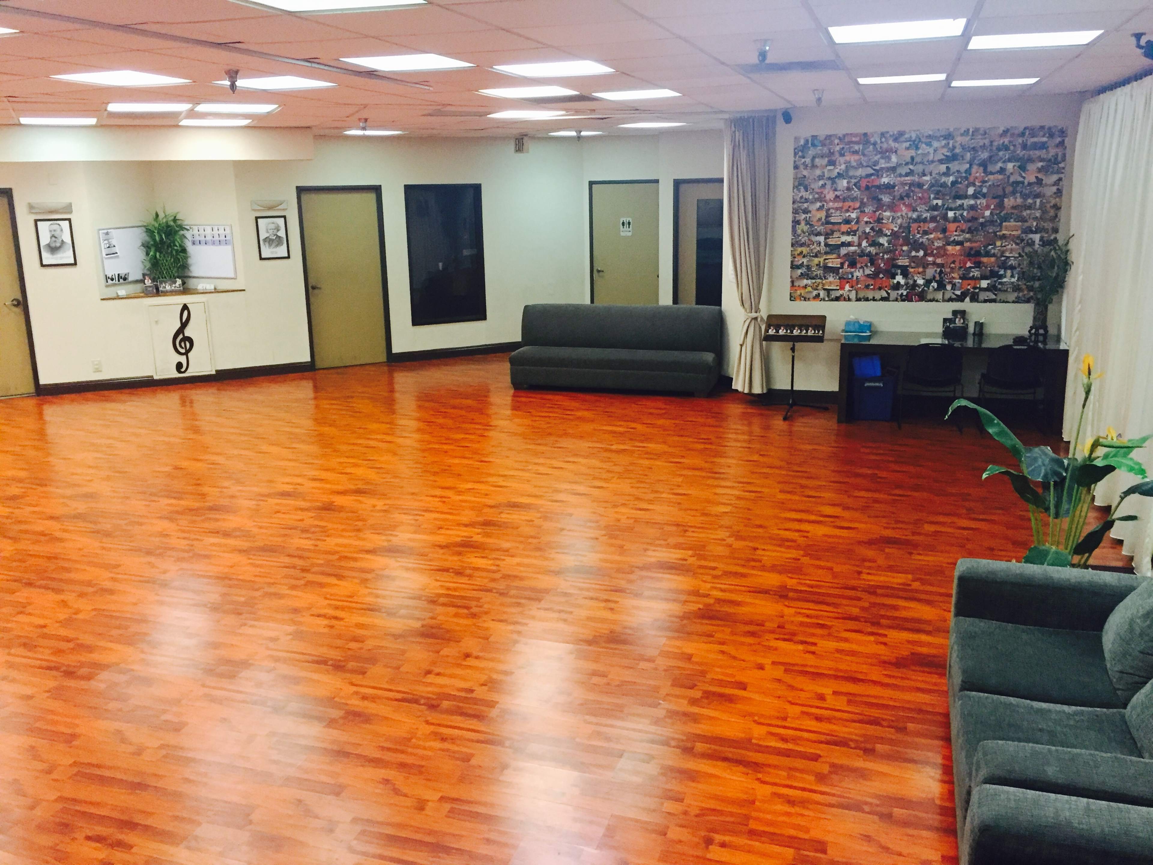 The image shows a spacious, wood-floored room with a few couches, walls adorned with framed art, and a small plant in the corner.