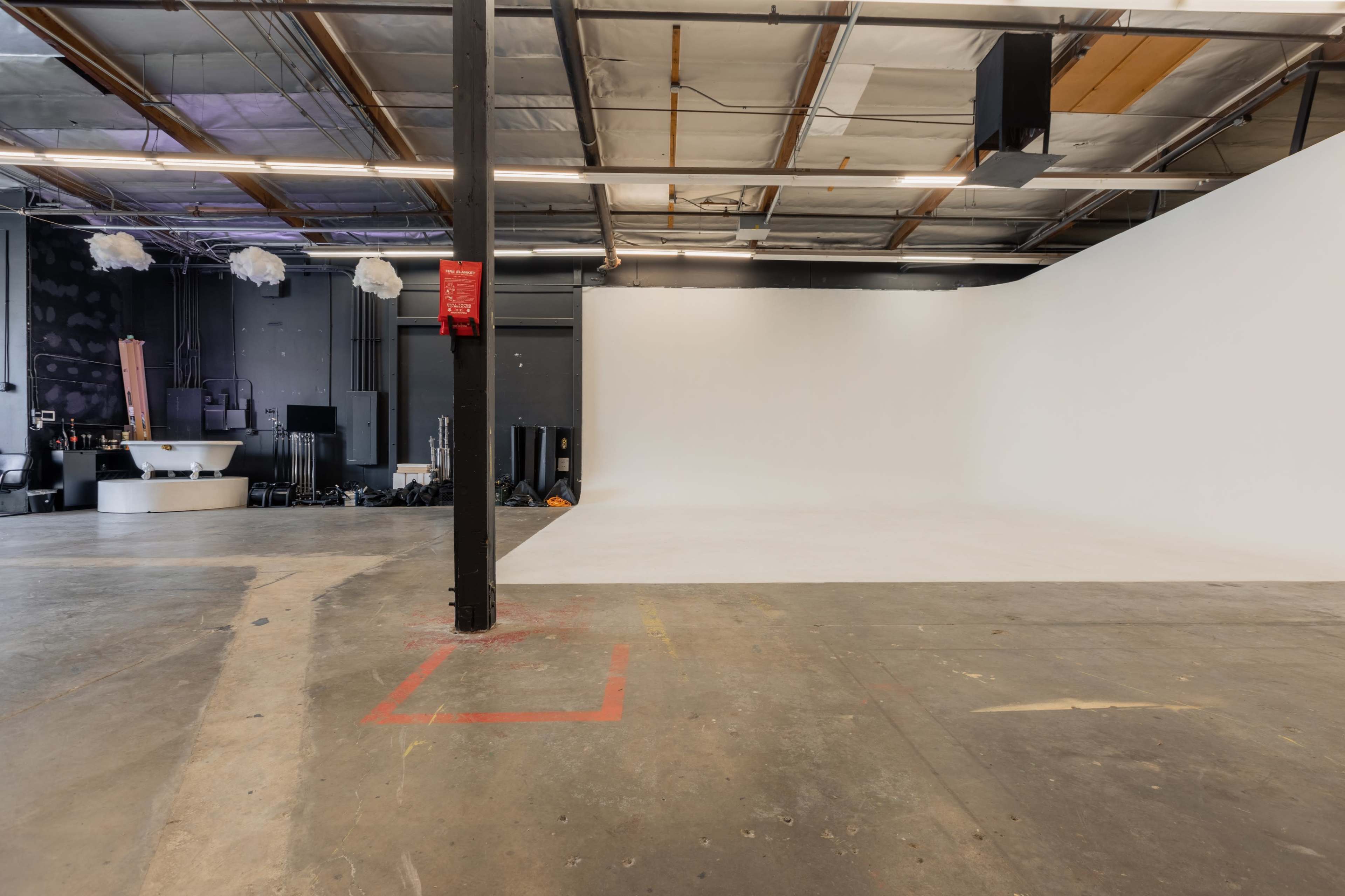 The image shows a spacious, empty studio with a white backdrop and concrete flooring, featuring a black vertical support beam in the foreground.