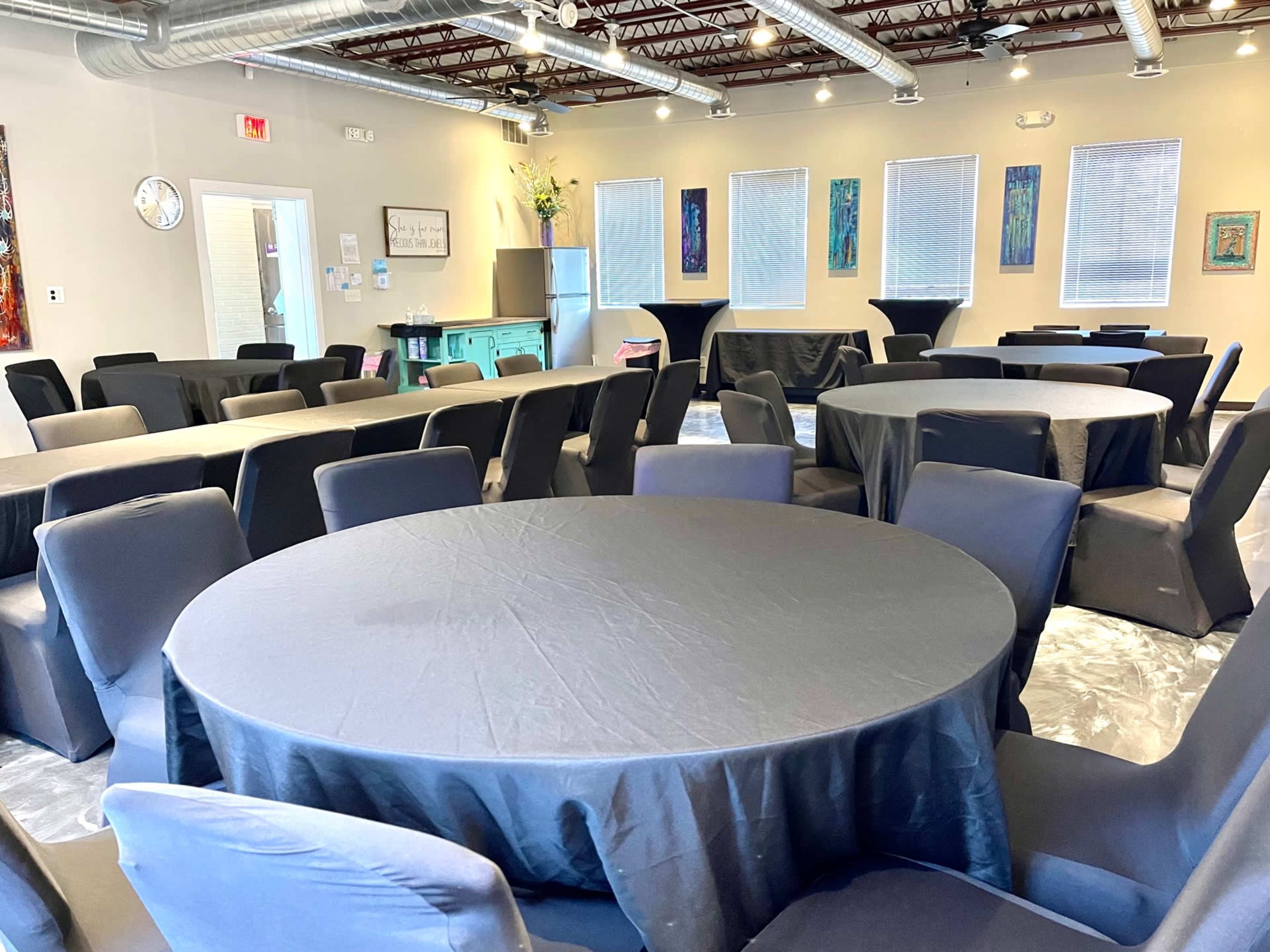 A spacious event room features several round tables covered with black tablecloths, surrounded by dark chairs.