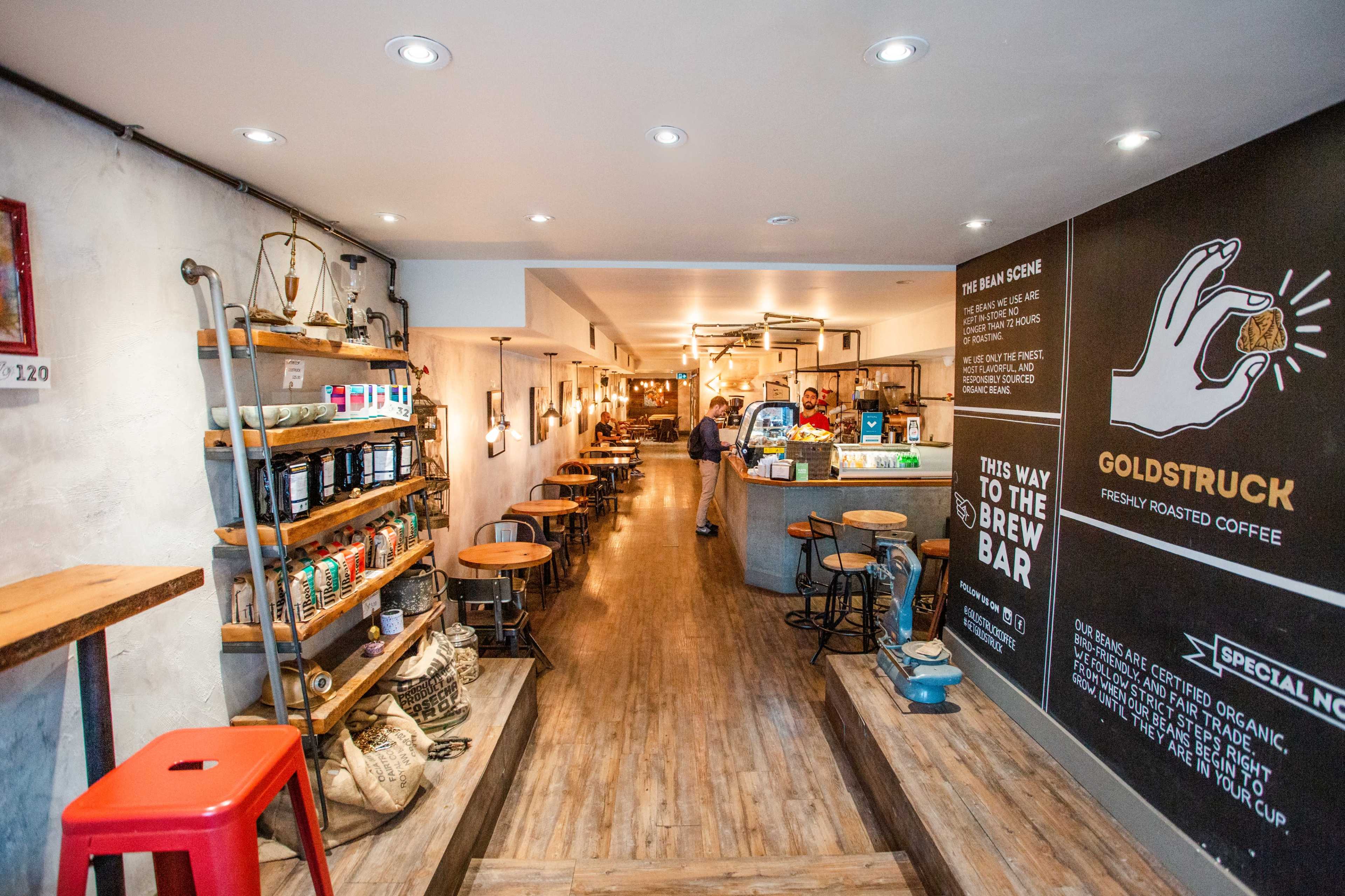 The image shows a modern coffee shop interior with wooden floors, shelves stocked with coffee products, and a counter at the back area.